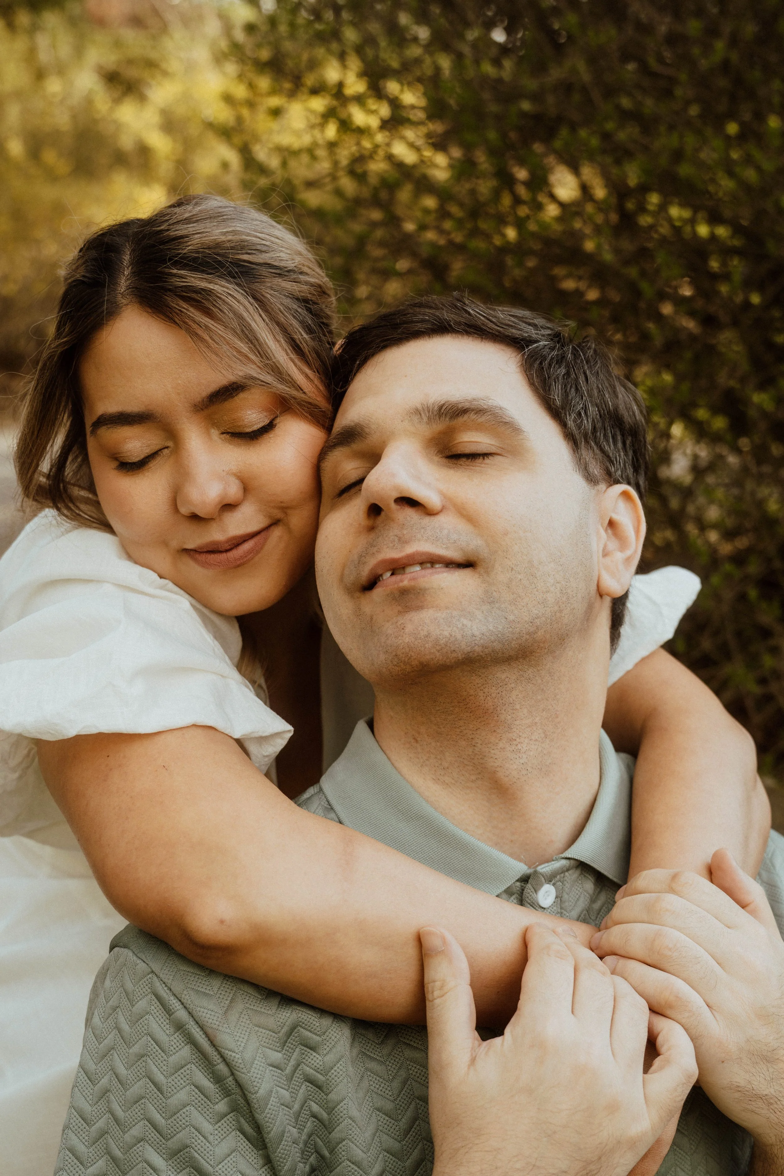 A couple hugging and smiling with eyes closed outdoors during daytime.