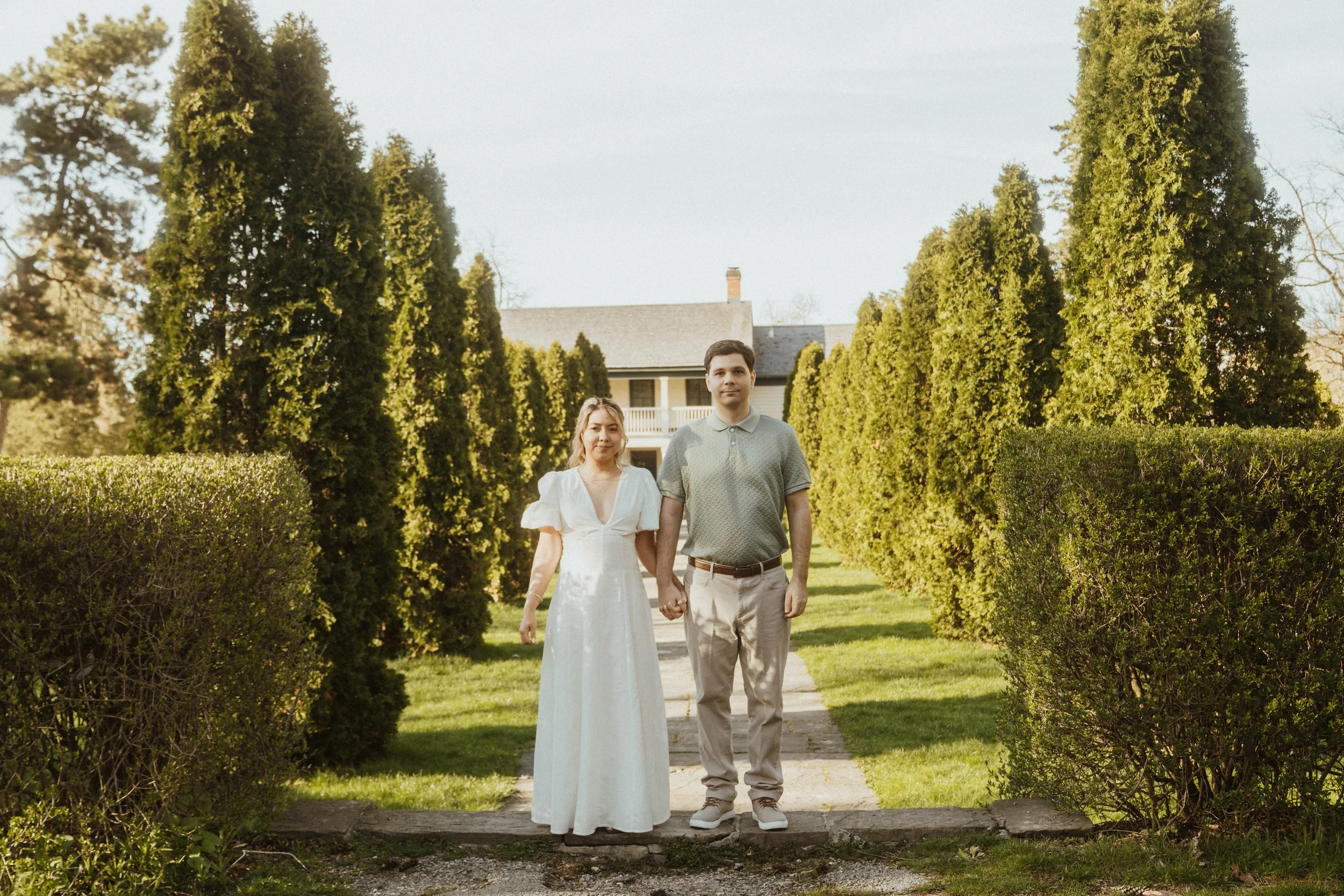 A young couple holding hands walking on a stone path between neatly trimmed hedges, with a house and tall trees in the background, during daytime.