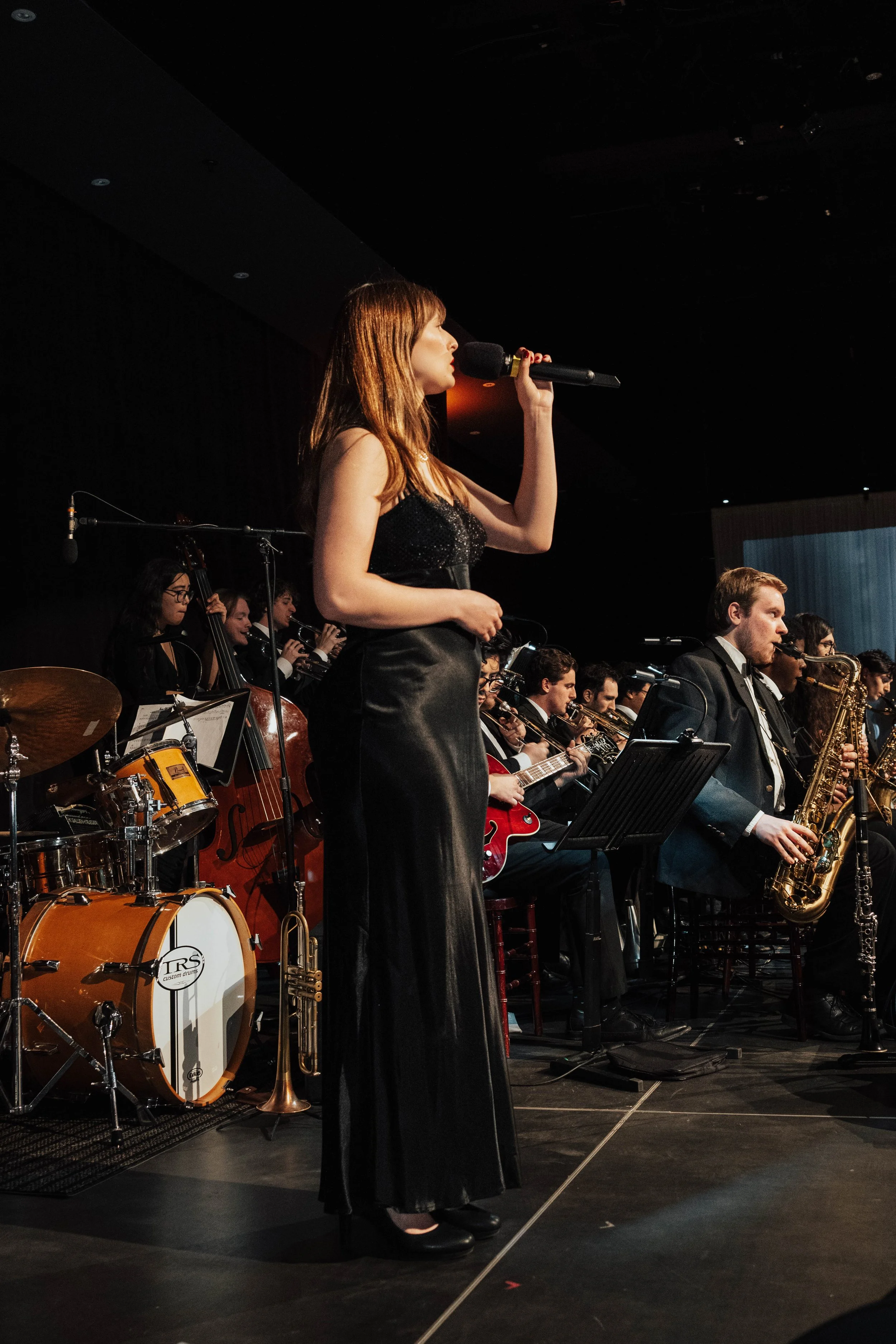 A woman singing into a microphone on stage with a band of musicians playing instruments behind her.