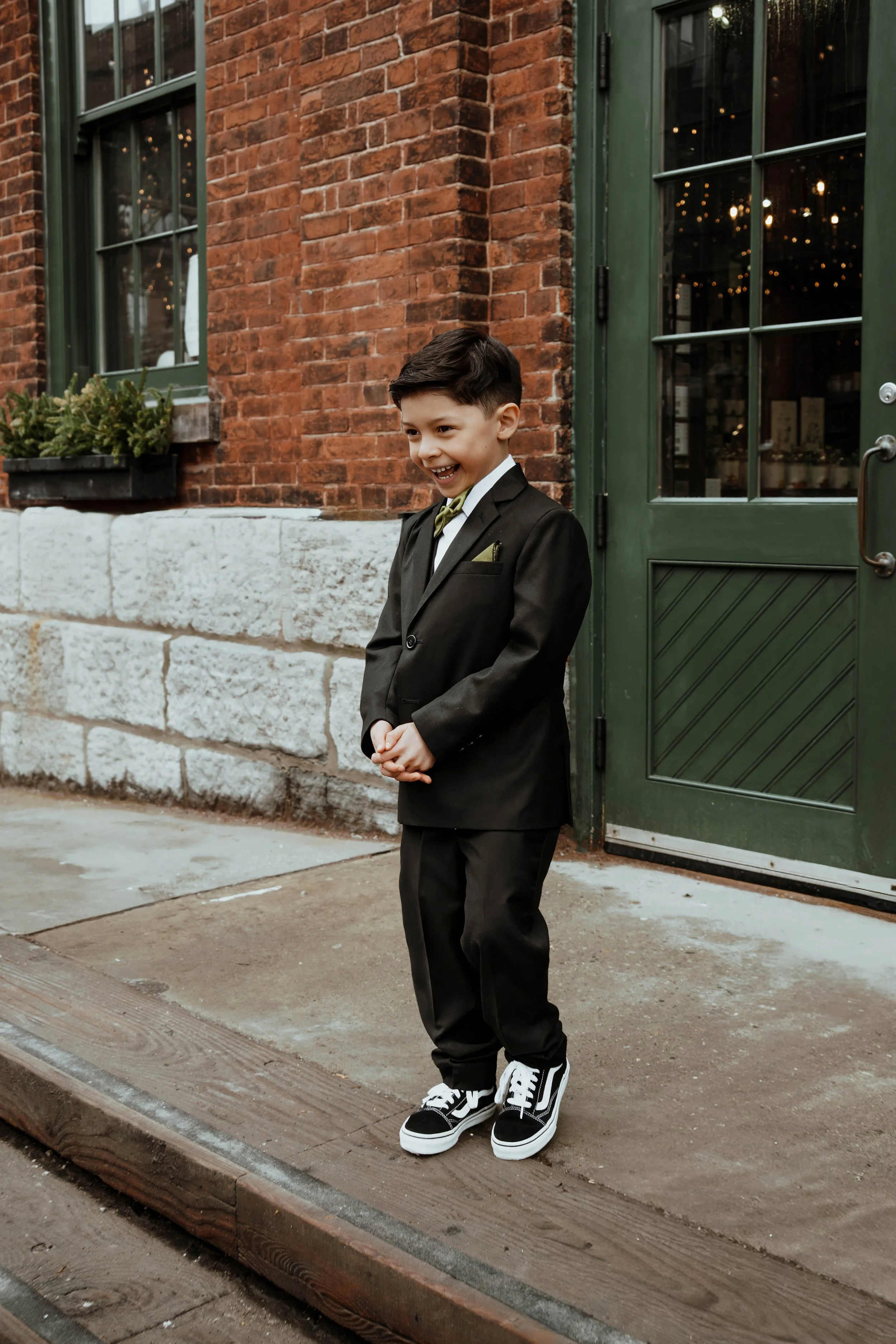 A young boy in a black suit, white shirt, and green bow tie, standing outside near a brick building with green door and window frames, smiling.