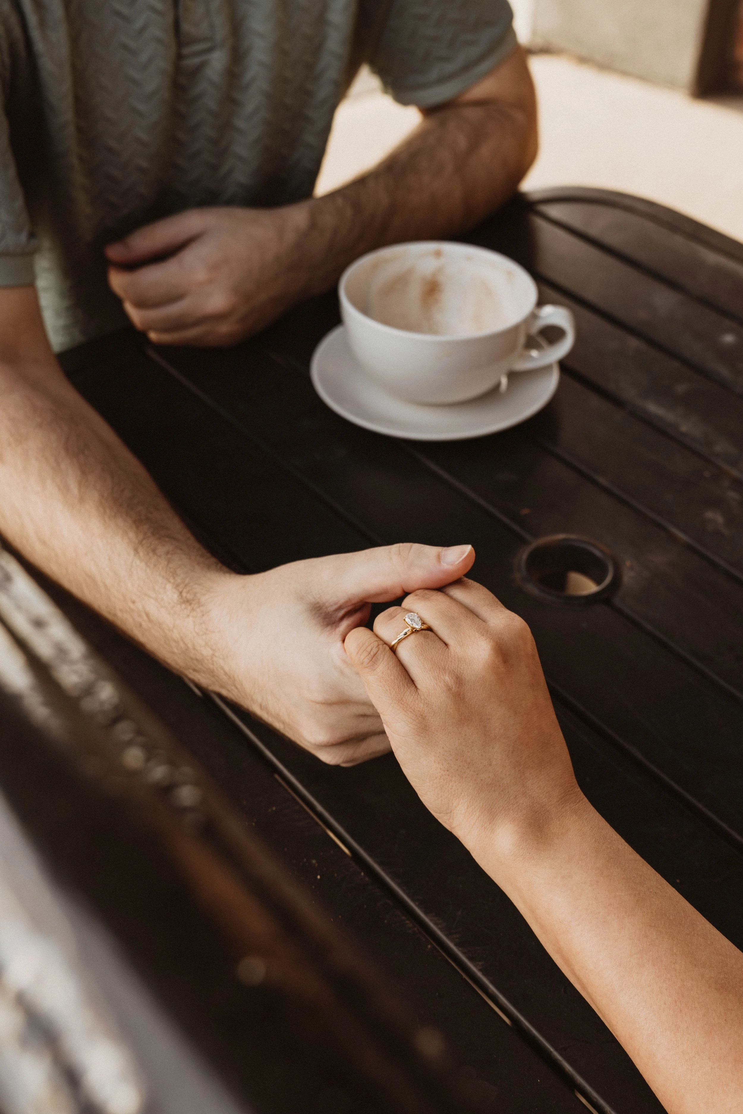 A woman with a manicured hand wearing a ring holds the hand of a man with light skin, sitting at a table with a cup of coffee that has frothy milk on top. A person's arm, wearing a gray short-sleeved shirt, is visible.