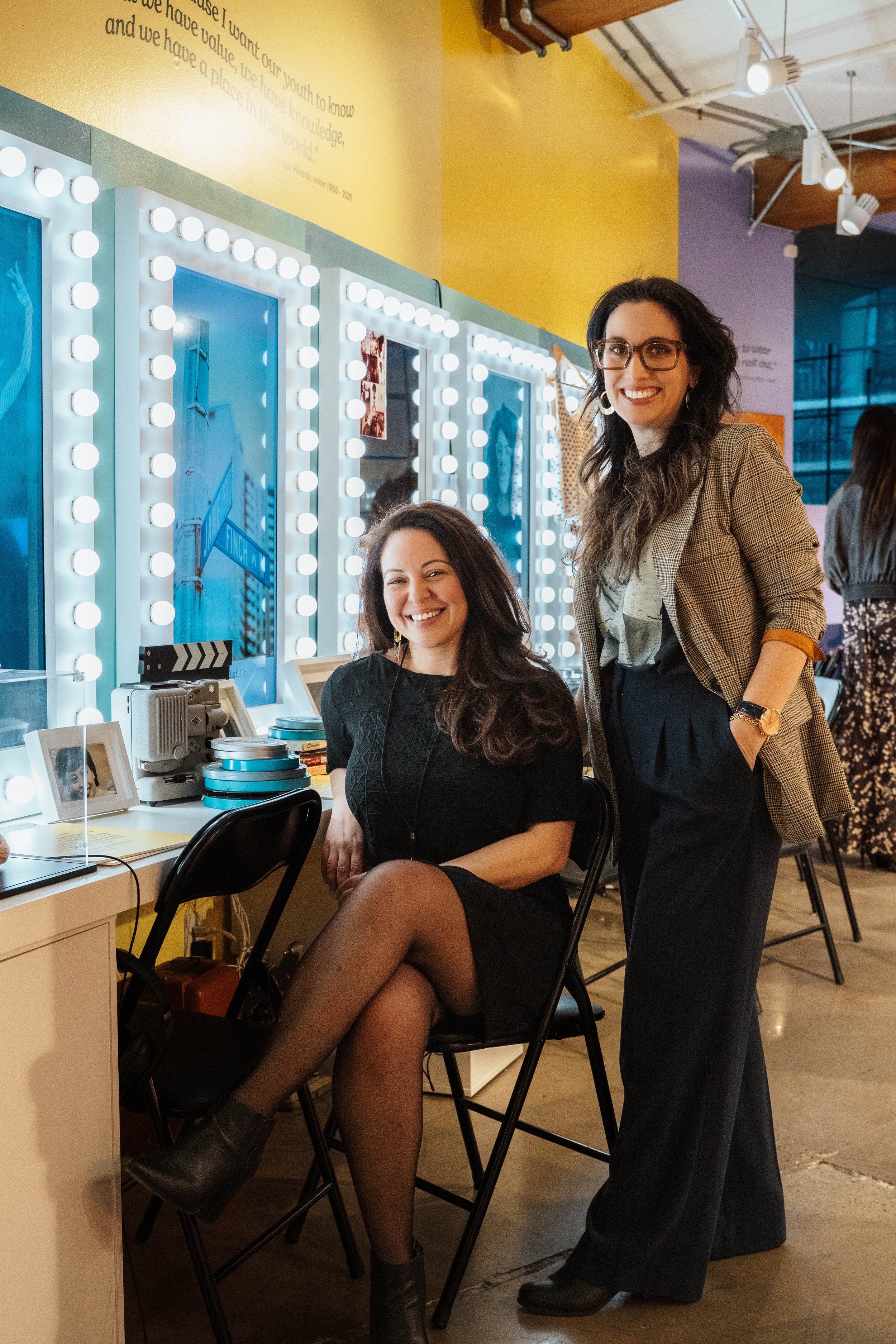 Two women in a brightly lit, colorful room with makeup mirrors. One woman is sitting on a chair, smiling, with crossed legs, wearing a black dress and stockings. The other woman is standing beside her, smiling, wearing glasses, a checked blazer, and 