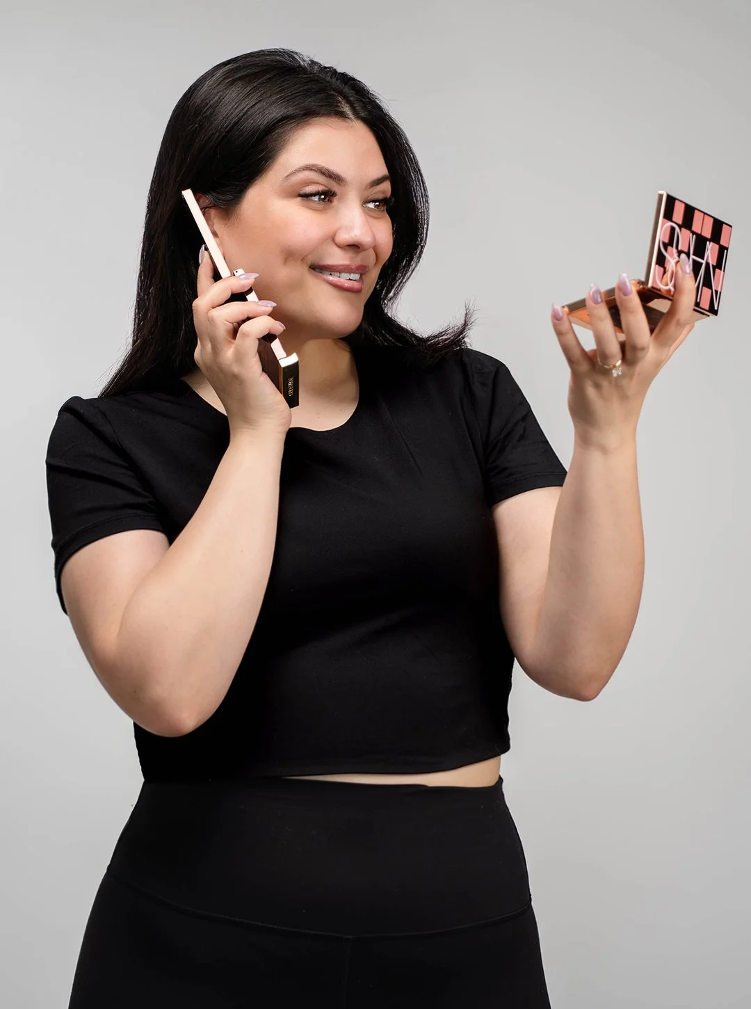 Woman with dark hair in black clothing applying makeup and holding a mirror.