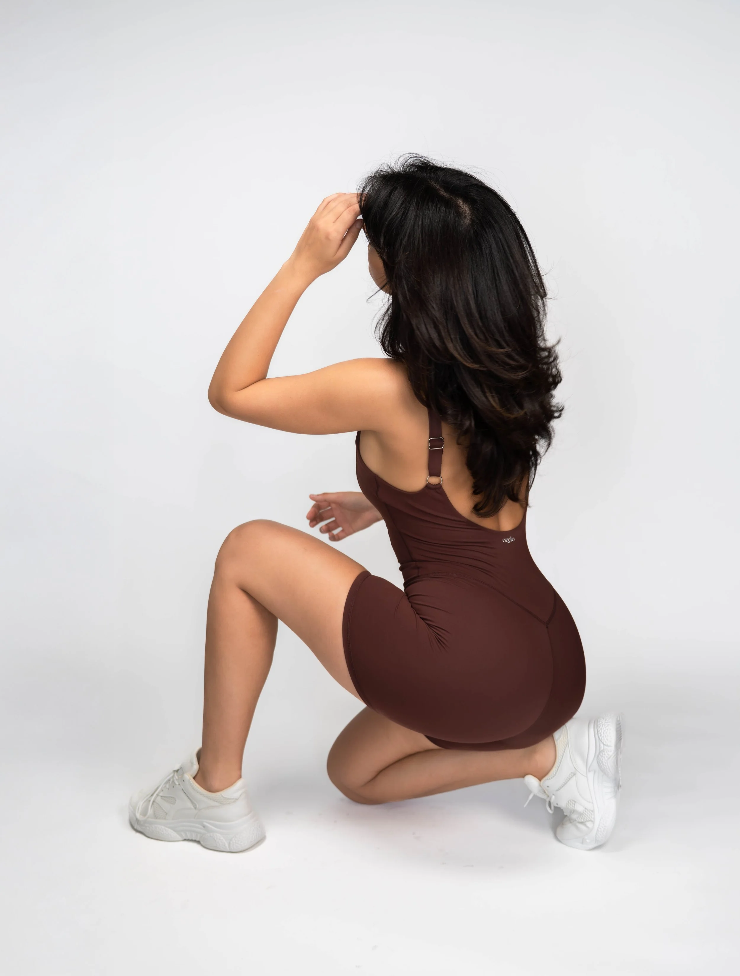 A woman with long dark hair wearing a brown sports outfit and white sneakers kneeling on the floor with one knee up, touching her head with her hand against a plain white background.