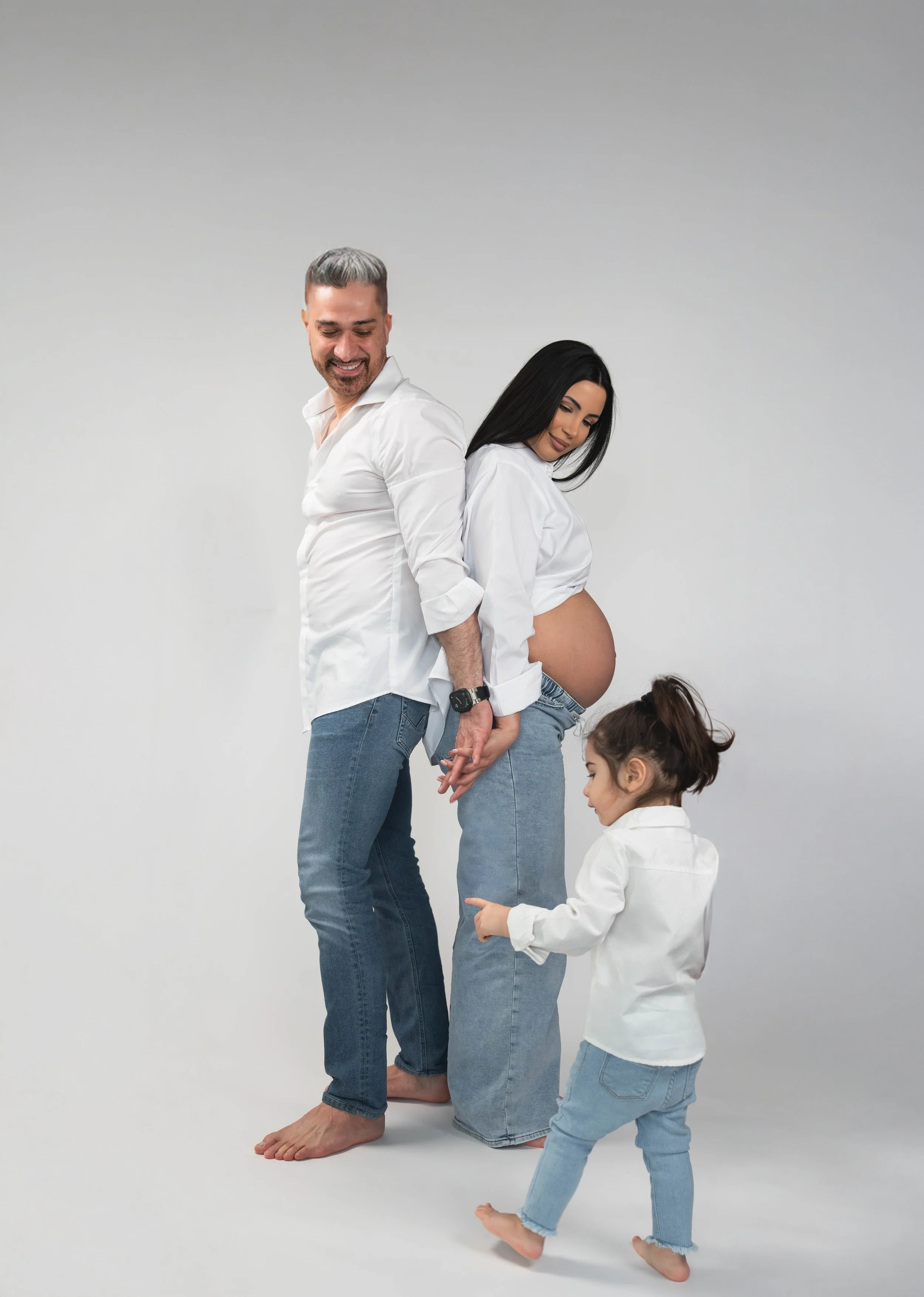 A family of four, including a pregnant woman, a man, a young girl, and a toddler girl, standing against a plain gray background. The man and pregnant woman are holding hands and standing back to back, smiling, with the man looking down and the woman 