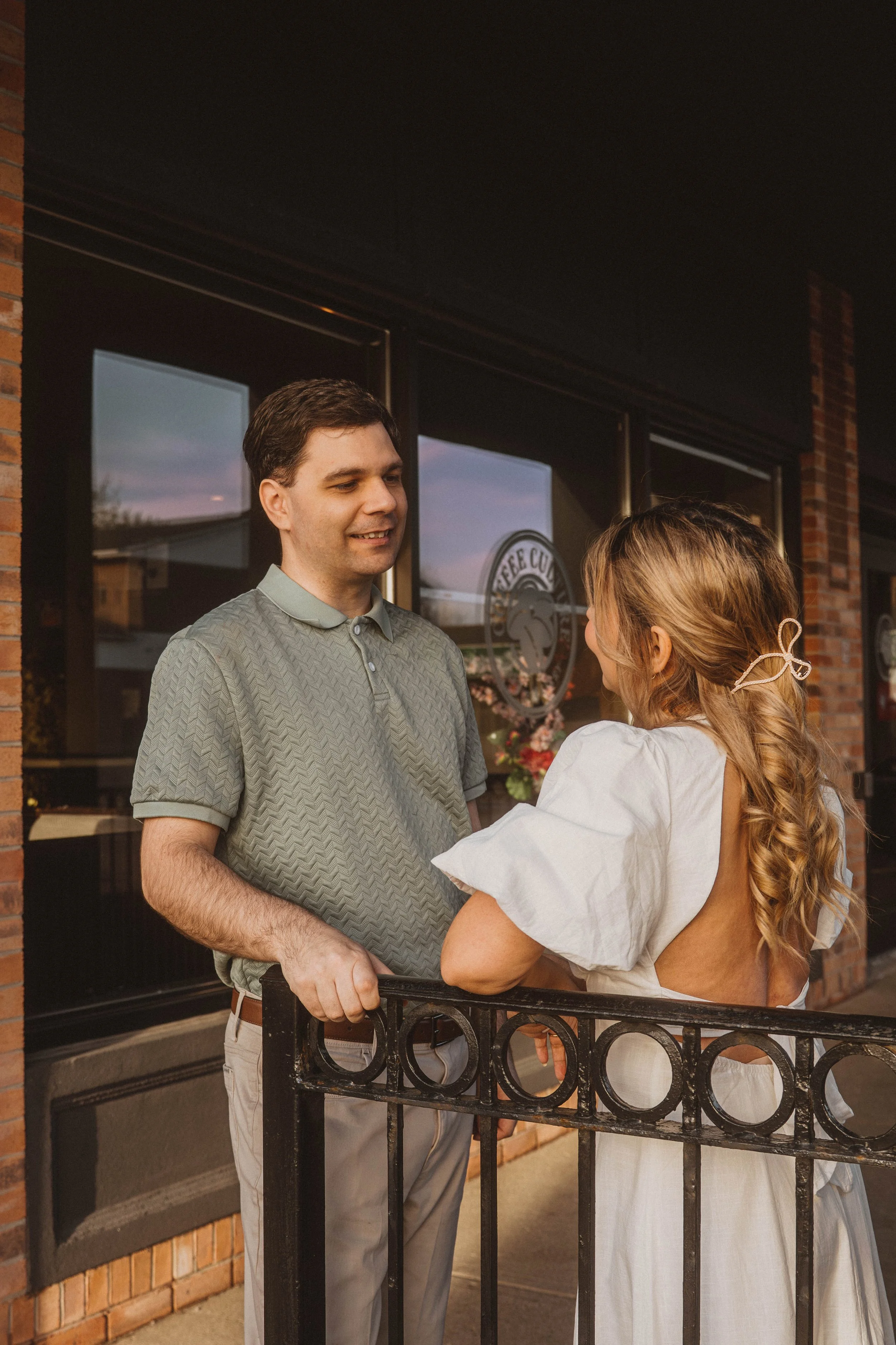 A man and woman are talking outside a building with large windows and brick walls. The man has dark hair and is wearing a light green polo shirt, while the woman has long, wavy, reddish-brown hair and is wearing a white dress. They are standing near 