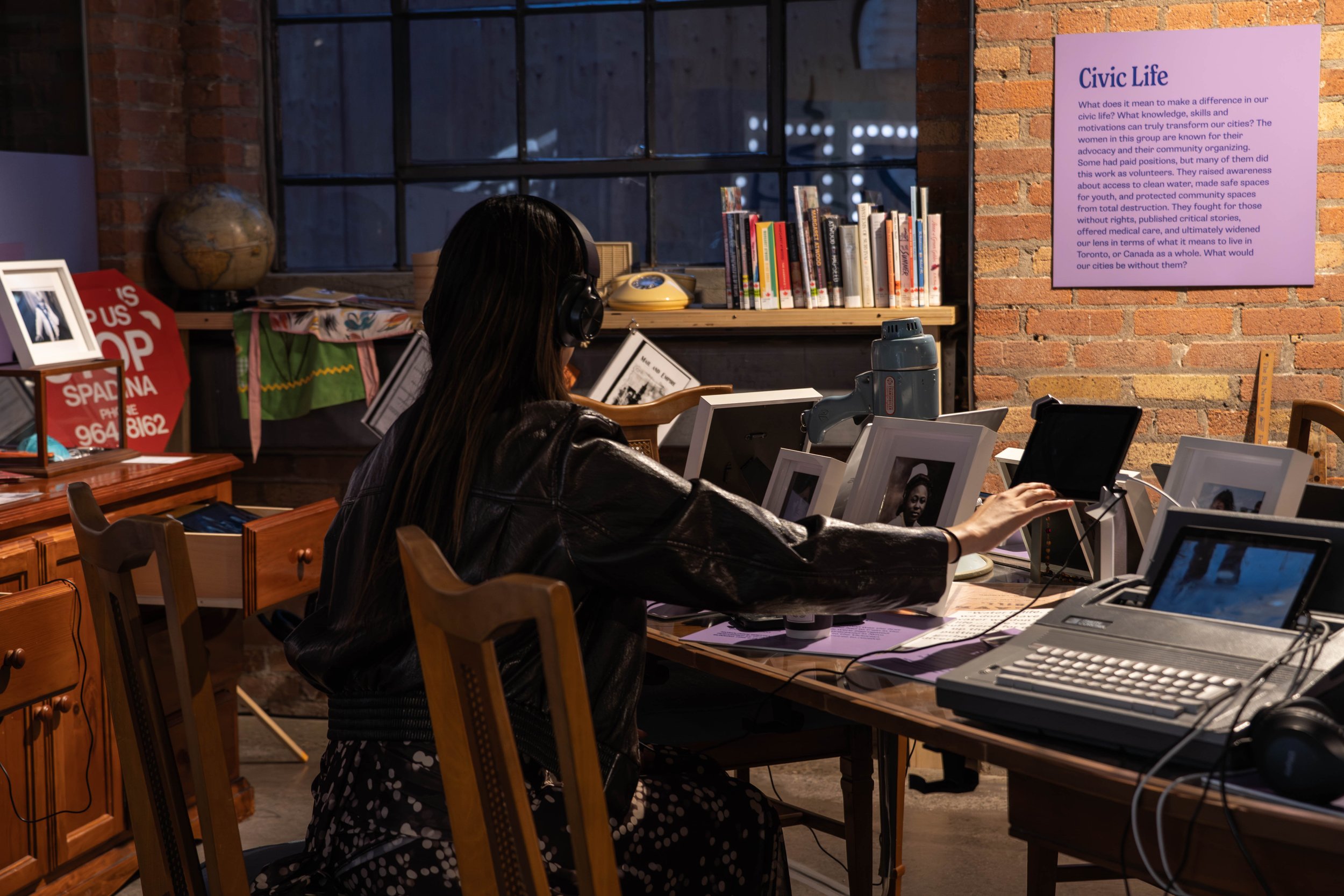 A woman with long dark hair wearing a black leather jacket and headphones sits at a cluttered office desk with multiple framed photos, a typewriter, and a computer as she reaches for a paper. Behind her is a brick wall with a purple infographic poste