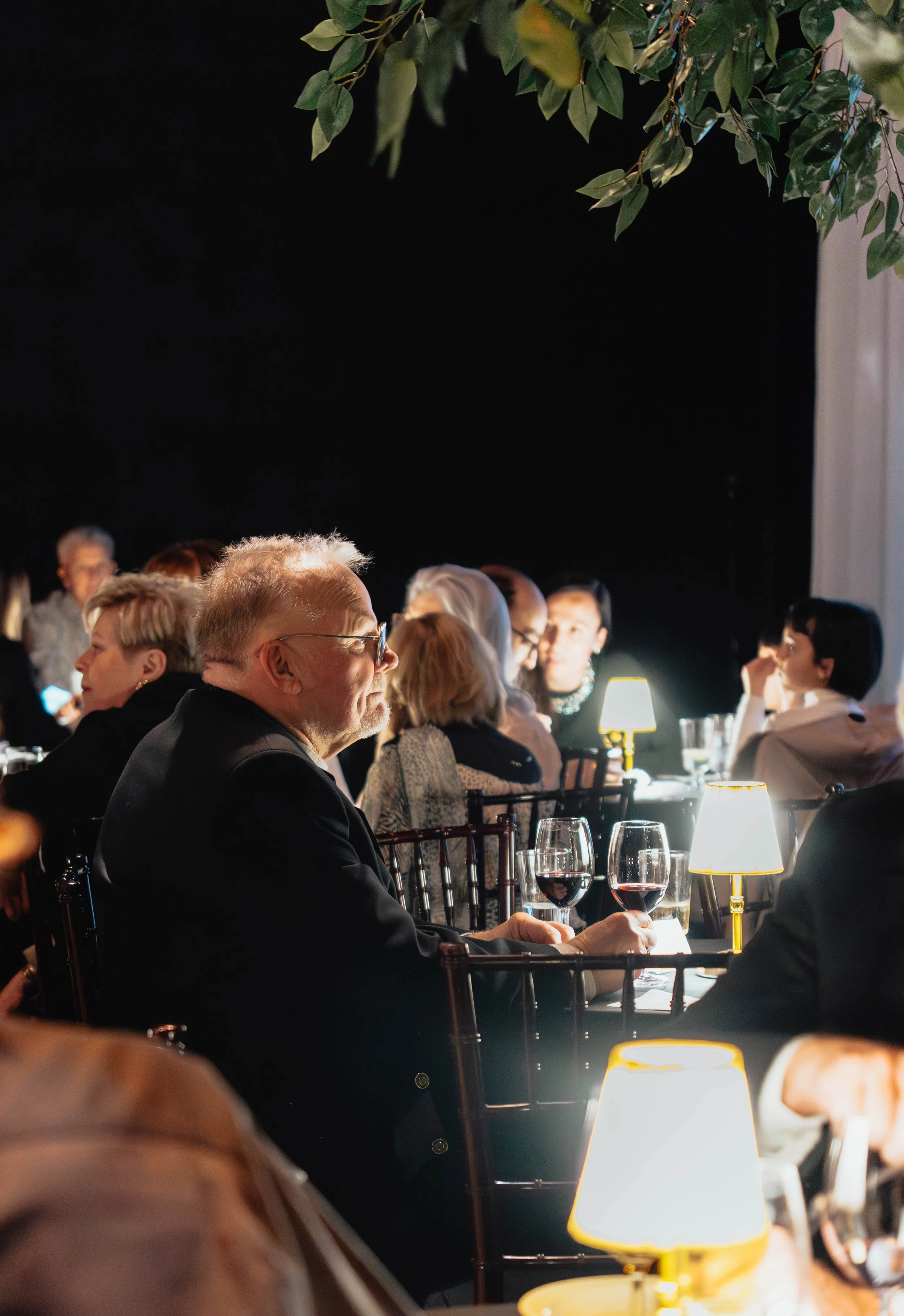 People seated at a formal dinner table, enjoying wine, with a dark background and small lamps providing warm lighting, at an indoor event.