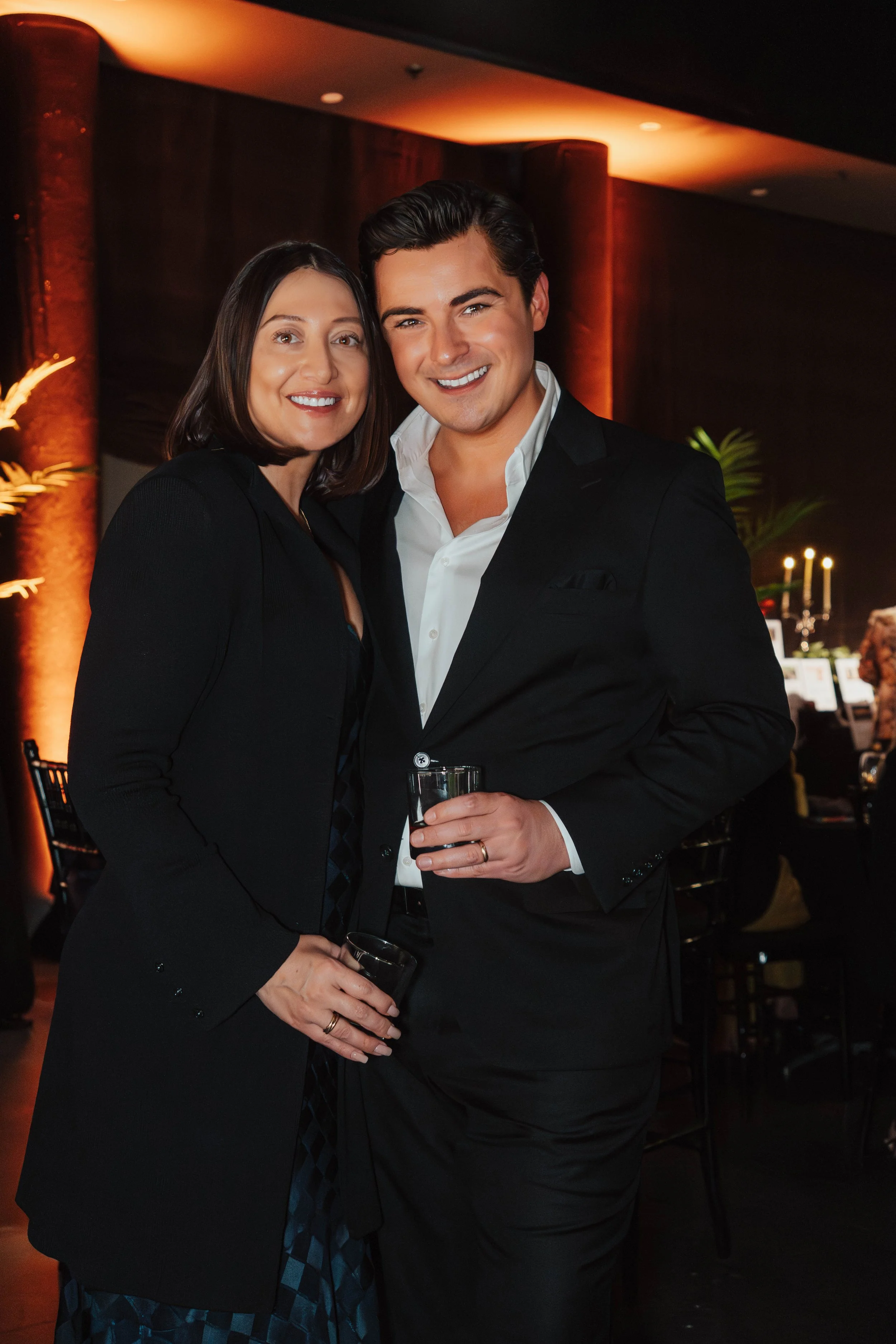 A smiling man and woman dressed in formal attire standing close together at a social event, holding drinks, with a warmly lit background.