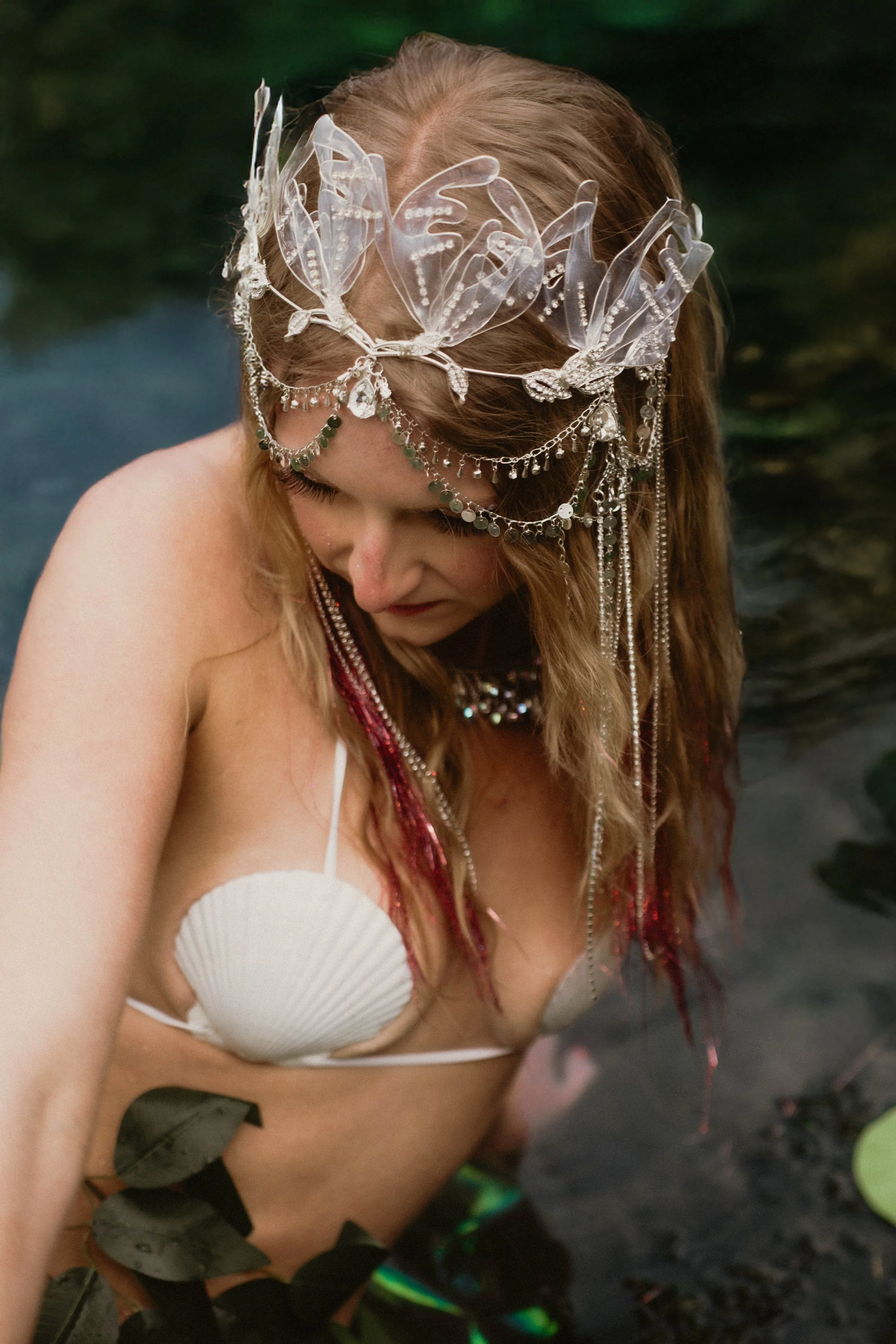 A woman with long red hair wearing a white bikini top and a decorative headpiece with clear butterflies and silver chains, standing near water surrounded by green plants.