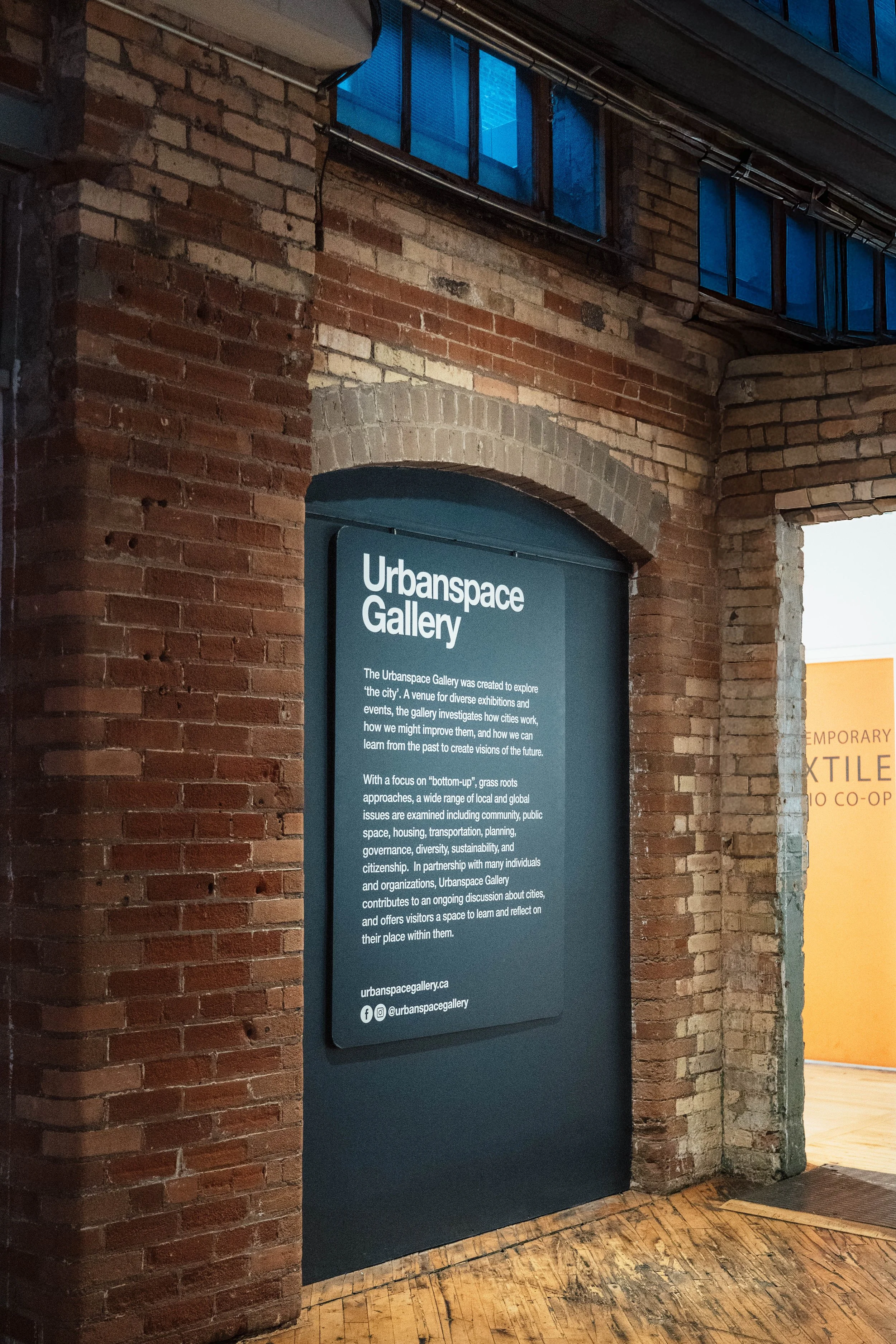 Signboard for Urbanspace Gallery explaining its purpose, focus, and partnership, mounted on a brick wall inside a building with wooden flooring, part of an industrial-style interior