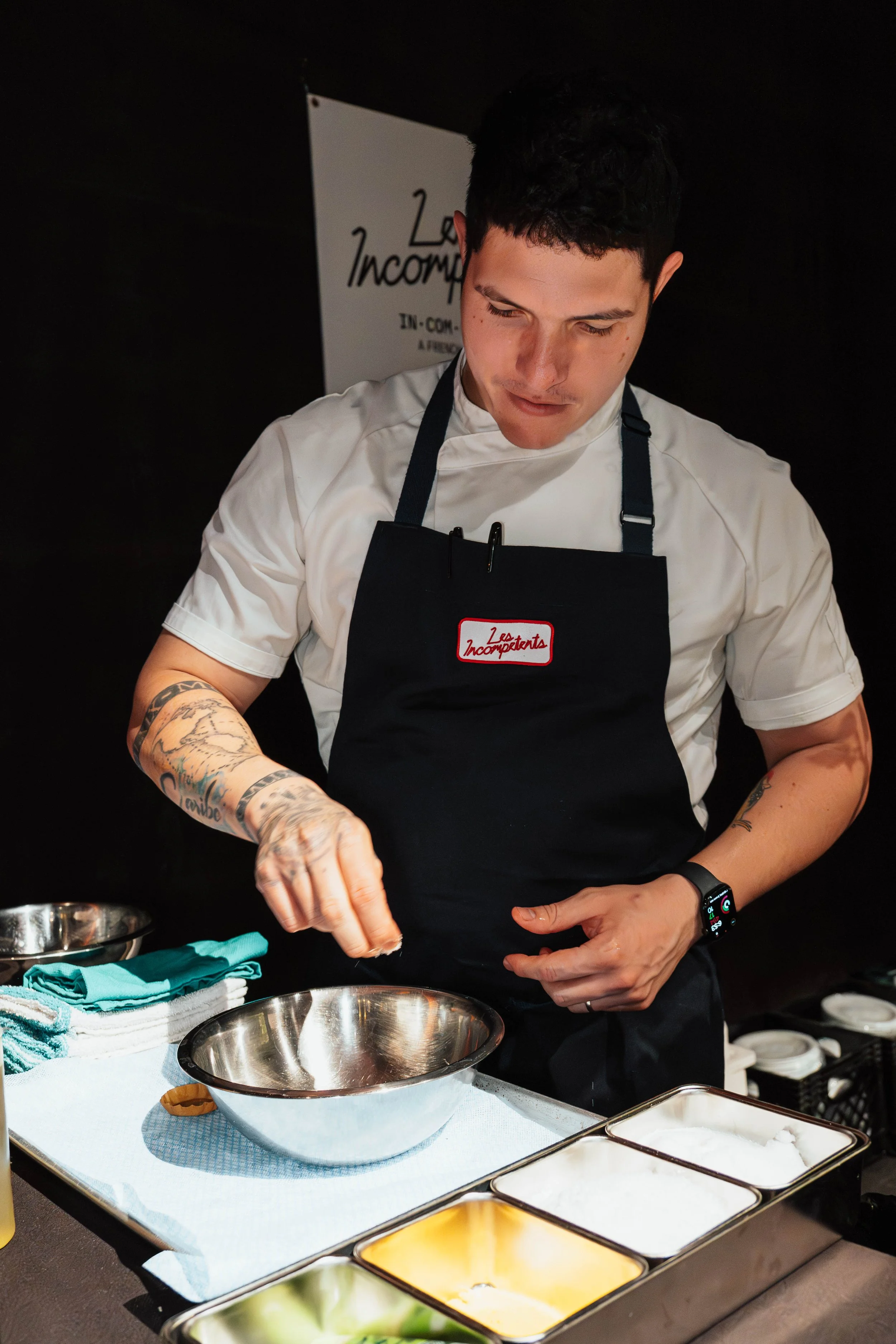 A man wearing a white chef's shirt and black apron with a red and white label is preparing food in a kitchen. He has tattoos on his right arm and is wearing a smartwatch. The countertop has bowls, towels, and ingredients, and there is a sign in the b