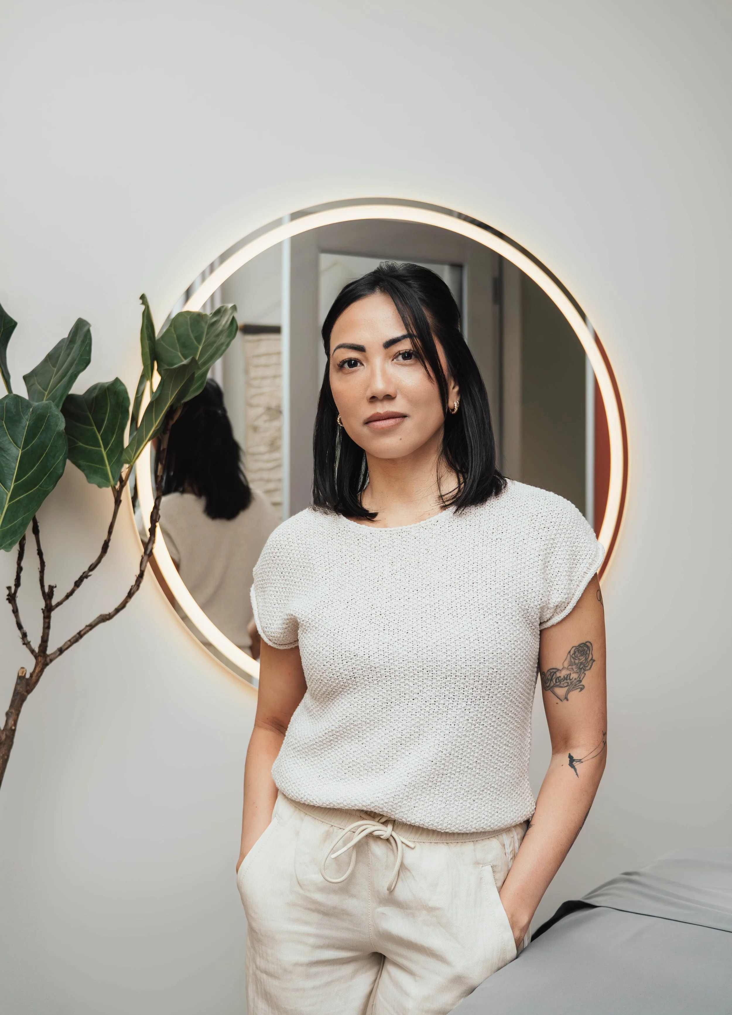 A woman with black hair and tattoos standing in front of a round mirror with a light around its edge, a plant next to her, and a bed partially visible in the foreground.