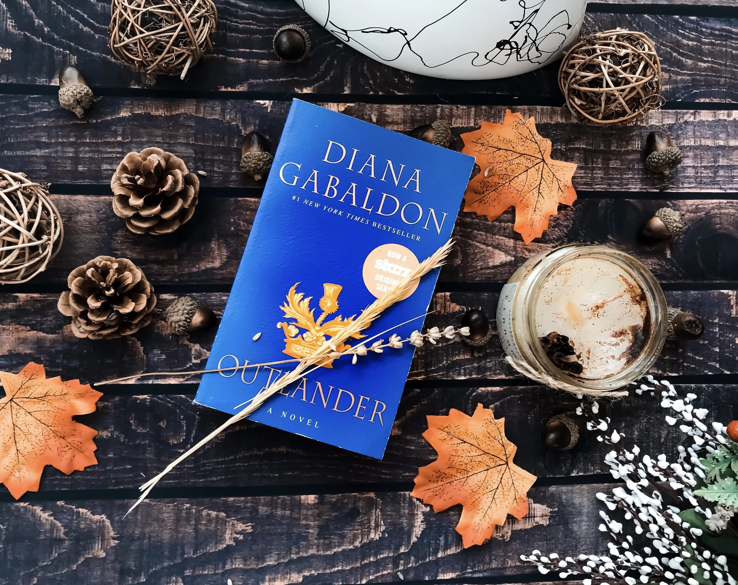 Book titled Outlander by Diana Gabaldon on a dark wooden surface surrounded by pinecones, acorns, orange autumn leaves, decorative wicker balls, a glass jar with remnants of a beverage and dried berries, and some white floral sprigs.