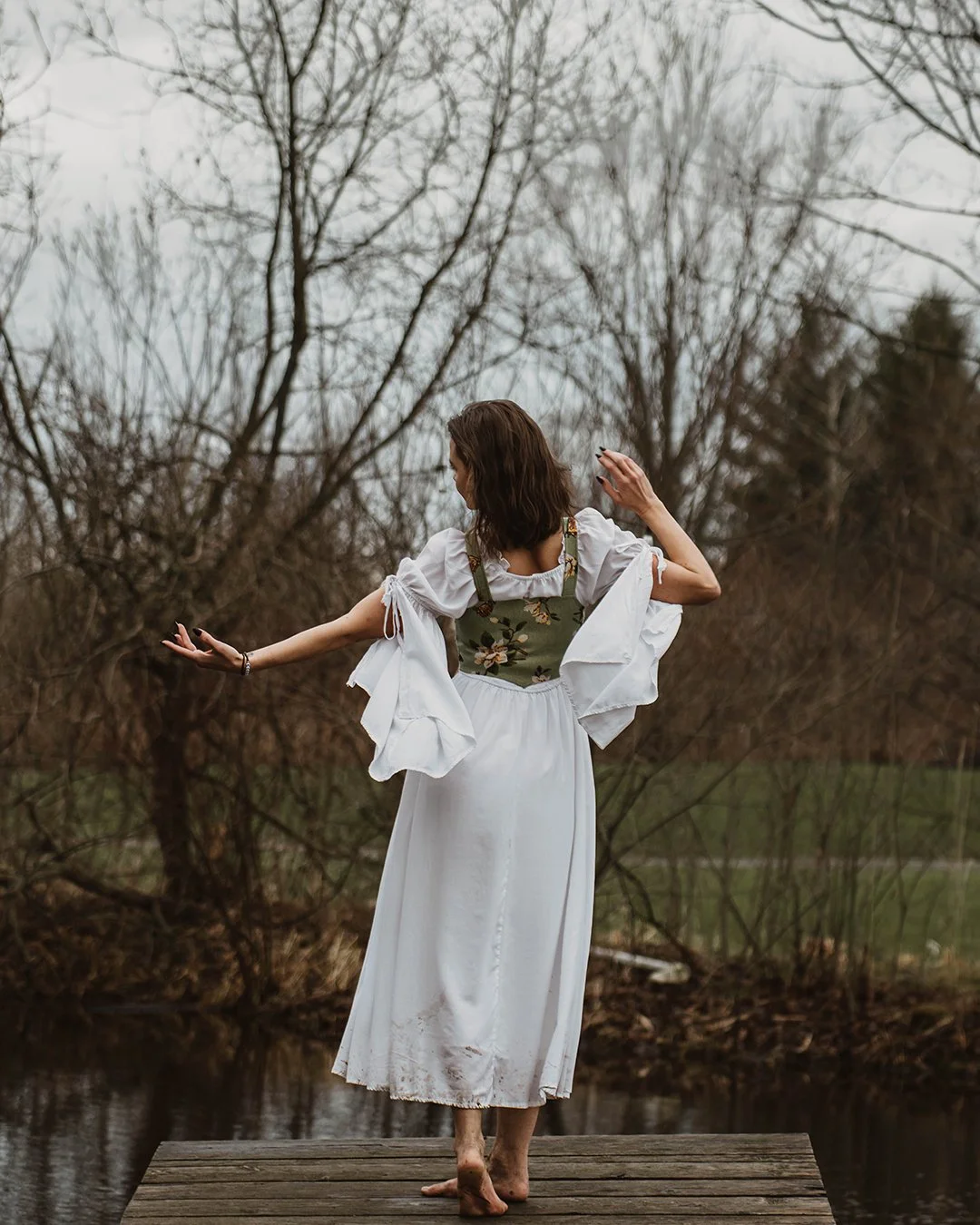 A woman in a white dress with puffed sleeves stands barefoot on a wooden dock by a pond, with her back to the camera, dancing or striking a pose amidst leafless trees and cloudy weather.