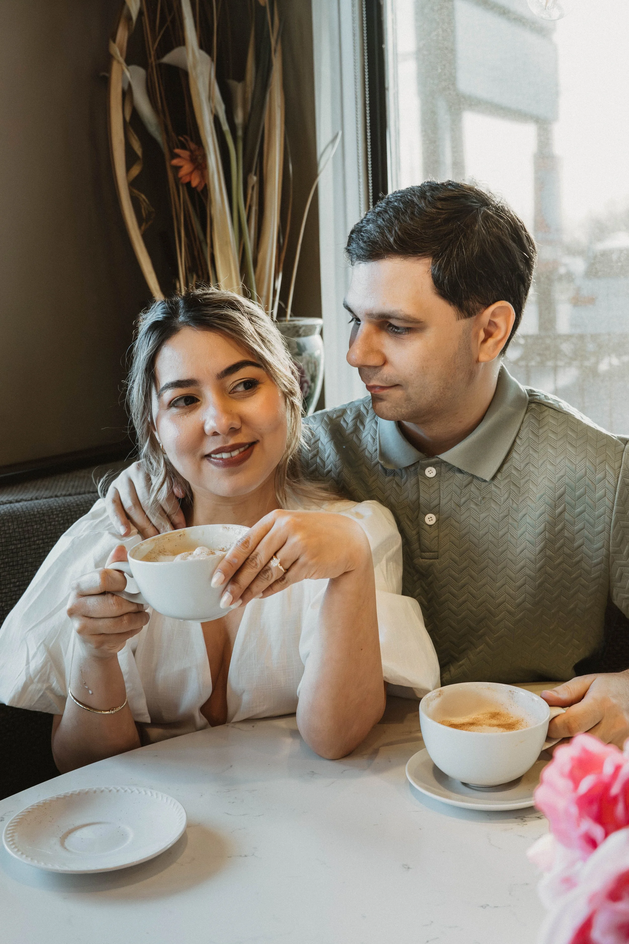 A woman with blonde hair and a man with dark hair sitting at a table, holding a cup of coffee with foam, in a cozy restaurant or cafe with natural light coming through a window. There are decorative plants behind them and a pink flower in the foregro