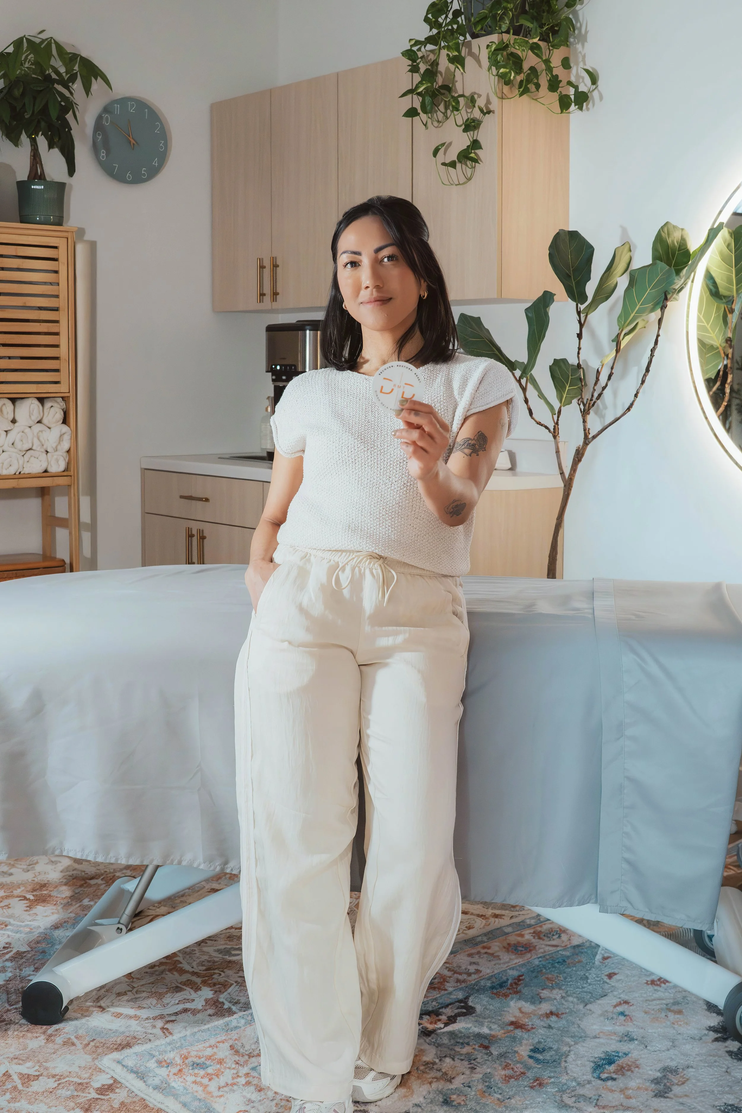 A woman standing and holding a small round object with a design on it, next to a massage table in a room with plants, wooden cabinets, and a clock on the wall.
