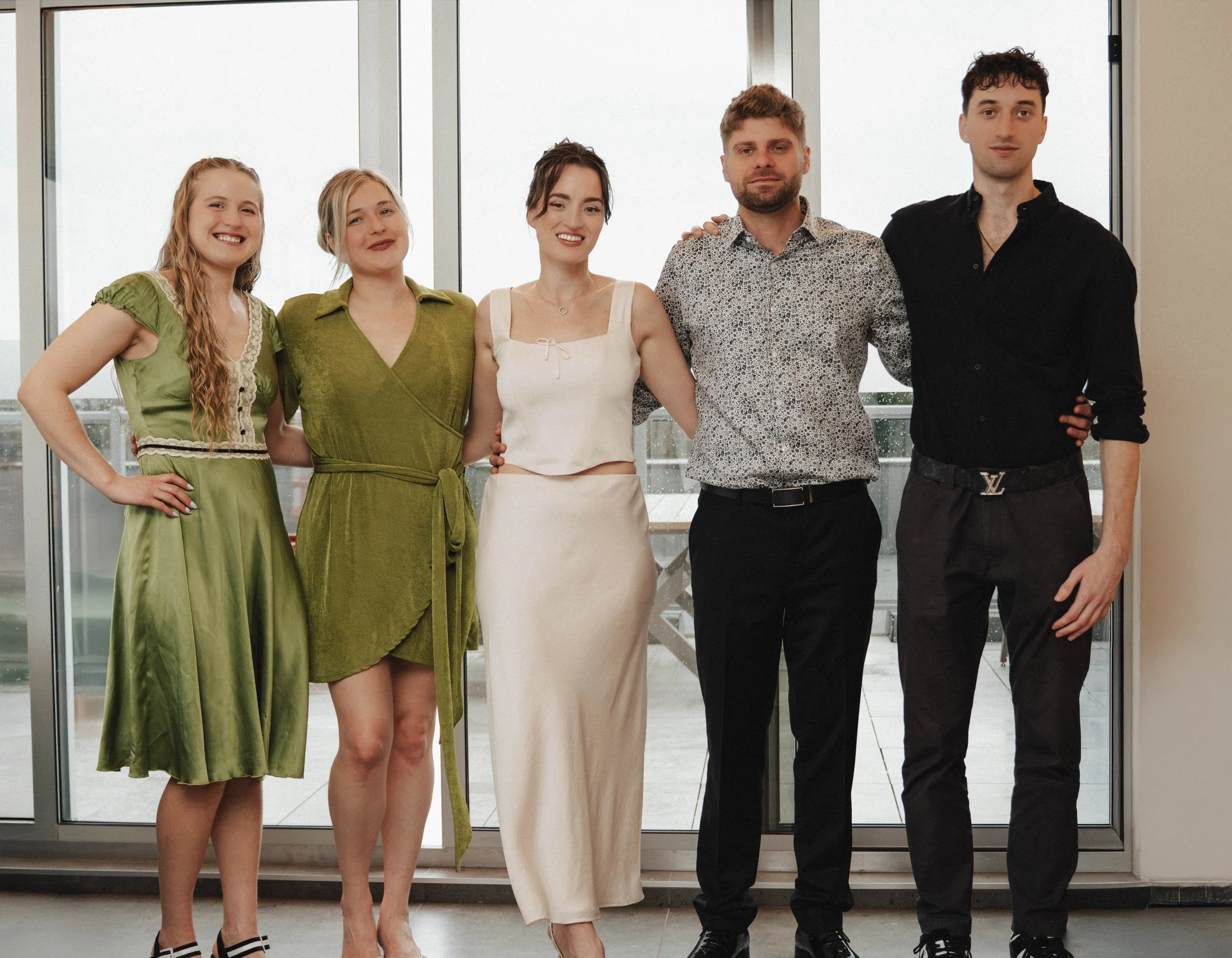 Five young adults standing together in front of a glass door, smiling at the camera. They are dressed in stylish, semi-formal clothing.