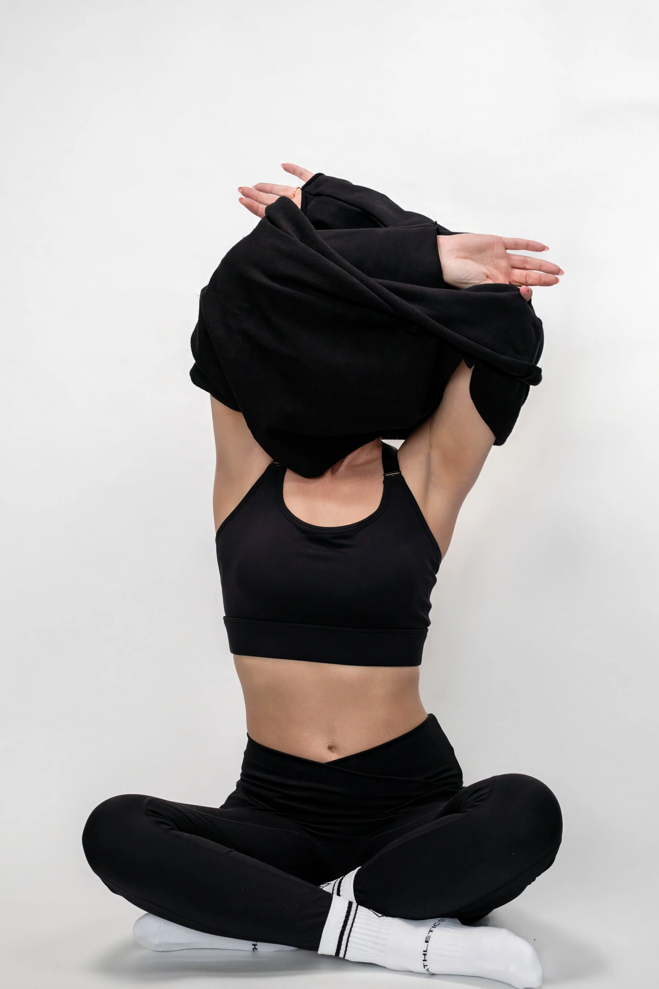Person sitting cross-legged in black athletic wear, lifting a black hoodie over her head against a plain white background.