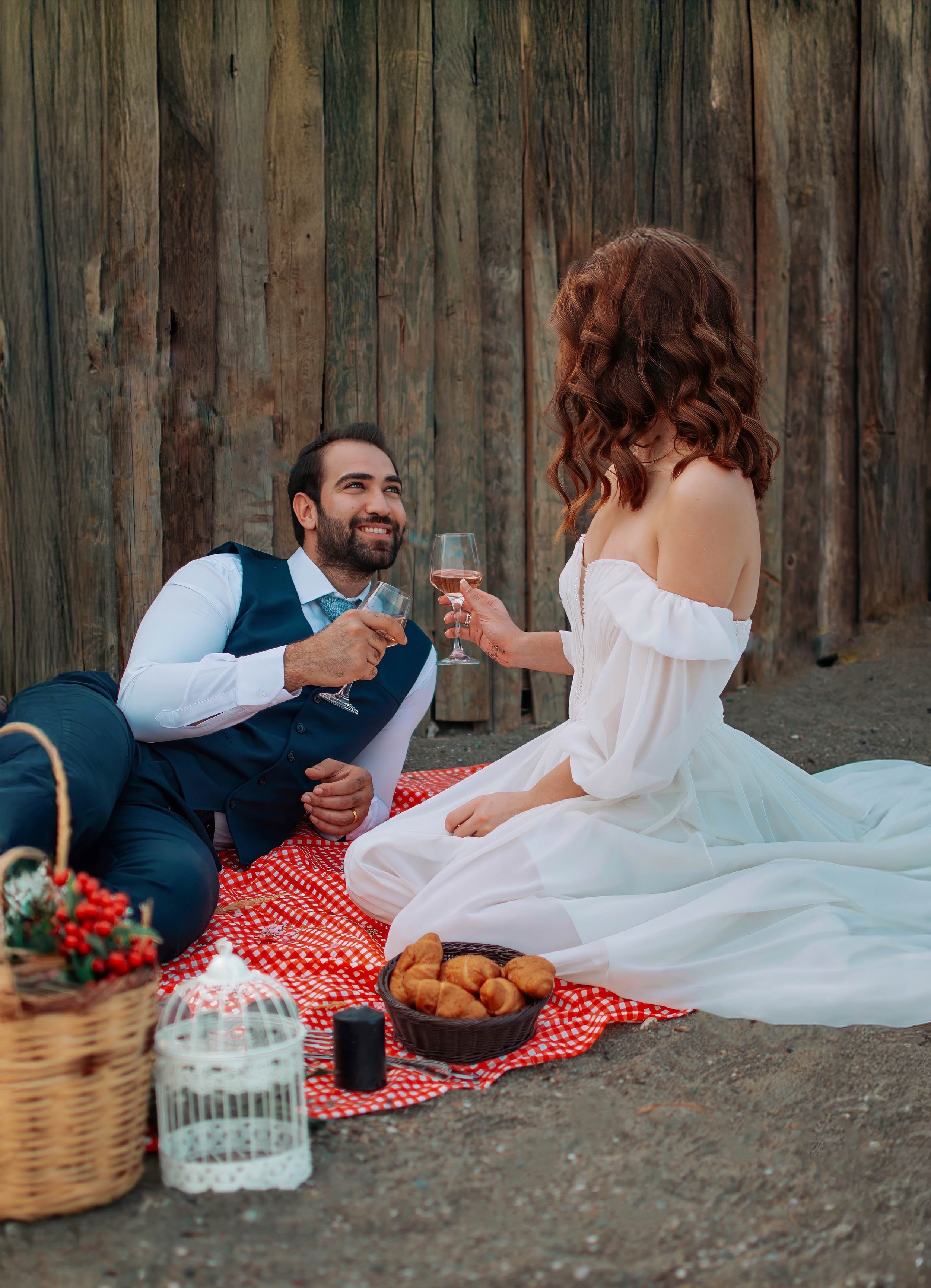 A couple having a picnic on the ground, with the man in a navy vest and tie and the woman in a white off-shoulder dress, both holding glasses of rosé wine, sitting on a red checkered blanket near a wooden fence.