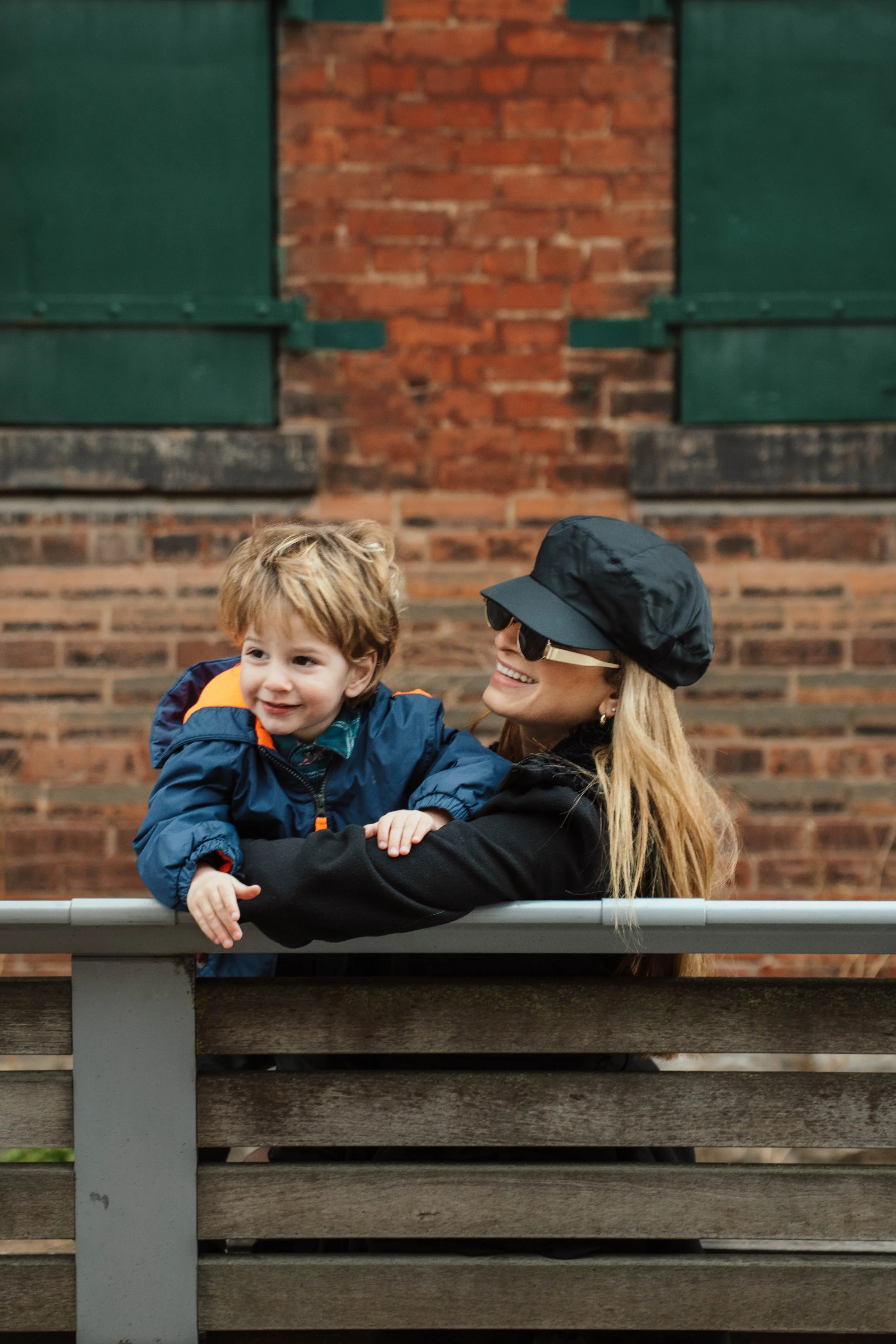 A woman with long blonde hair wearing sunglasses, a black hat, and a black jacket, holding a young boy with curly blonde hair, a blue jacket, and an orange hood, in front of an old brick wall with green shutters.