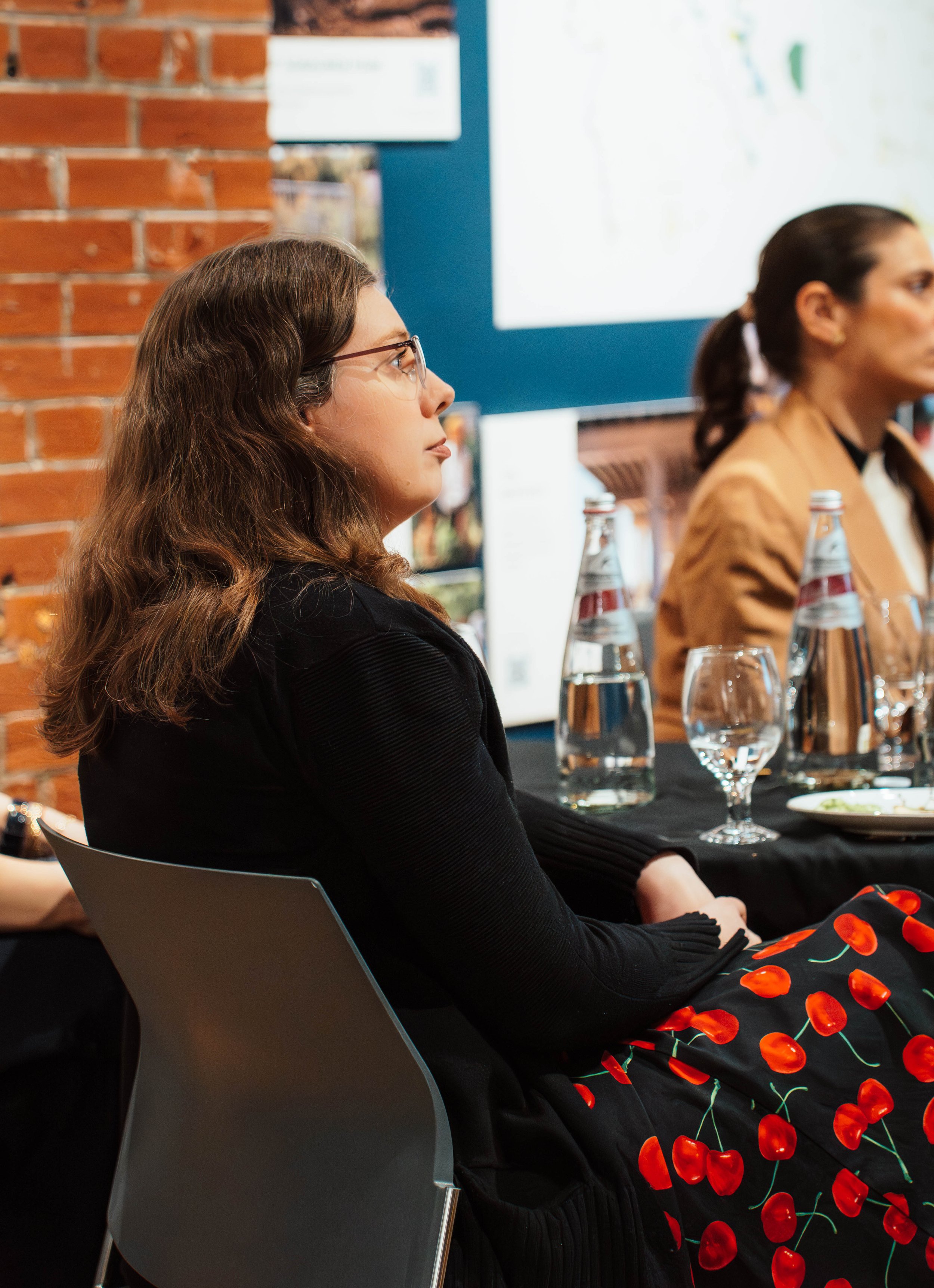 A woman with long brown hair and glasses, sitting at a table during a meeting or presentation, with bottled water and glasses in front of her, in a room with brick walls and a large screen.