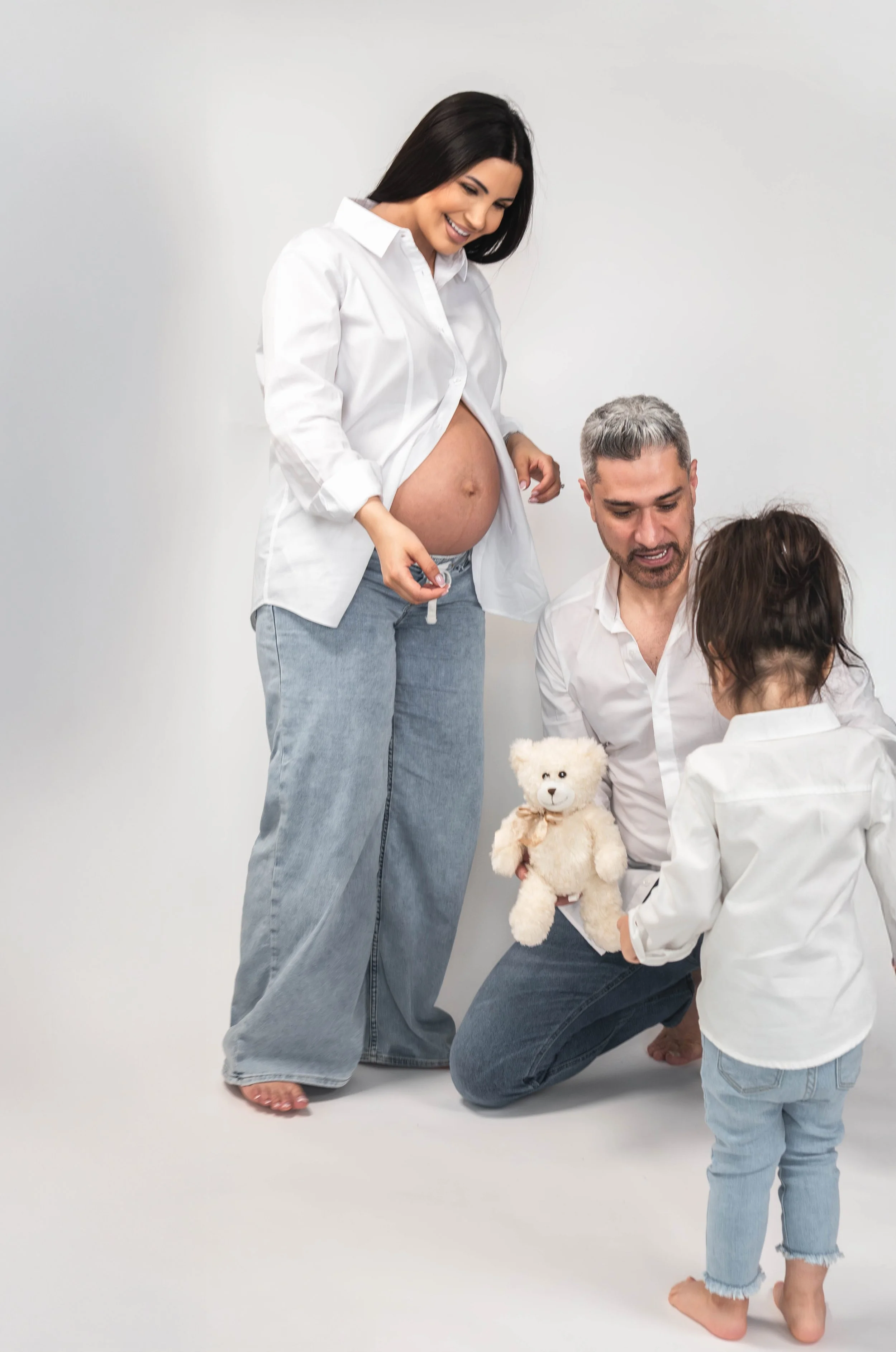 A pregnant woman in a white shirt and jeans smiling at her family, with her belly exposed. Her partner, kneeling, holds a teddy bear and looks at their child, with a woman and a child present in a light-colored studio setting.