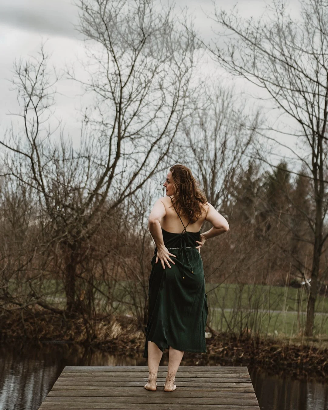 A woman in a black dress standing barefoot on a wooden dock by a body of water, with leafless trees in the background during overcast weather.