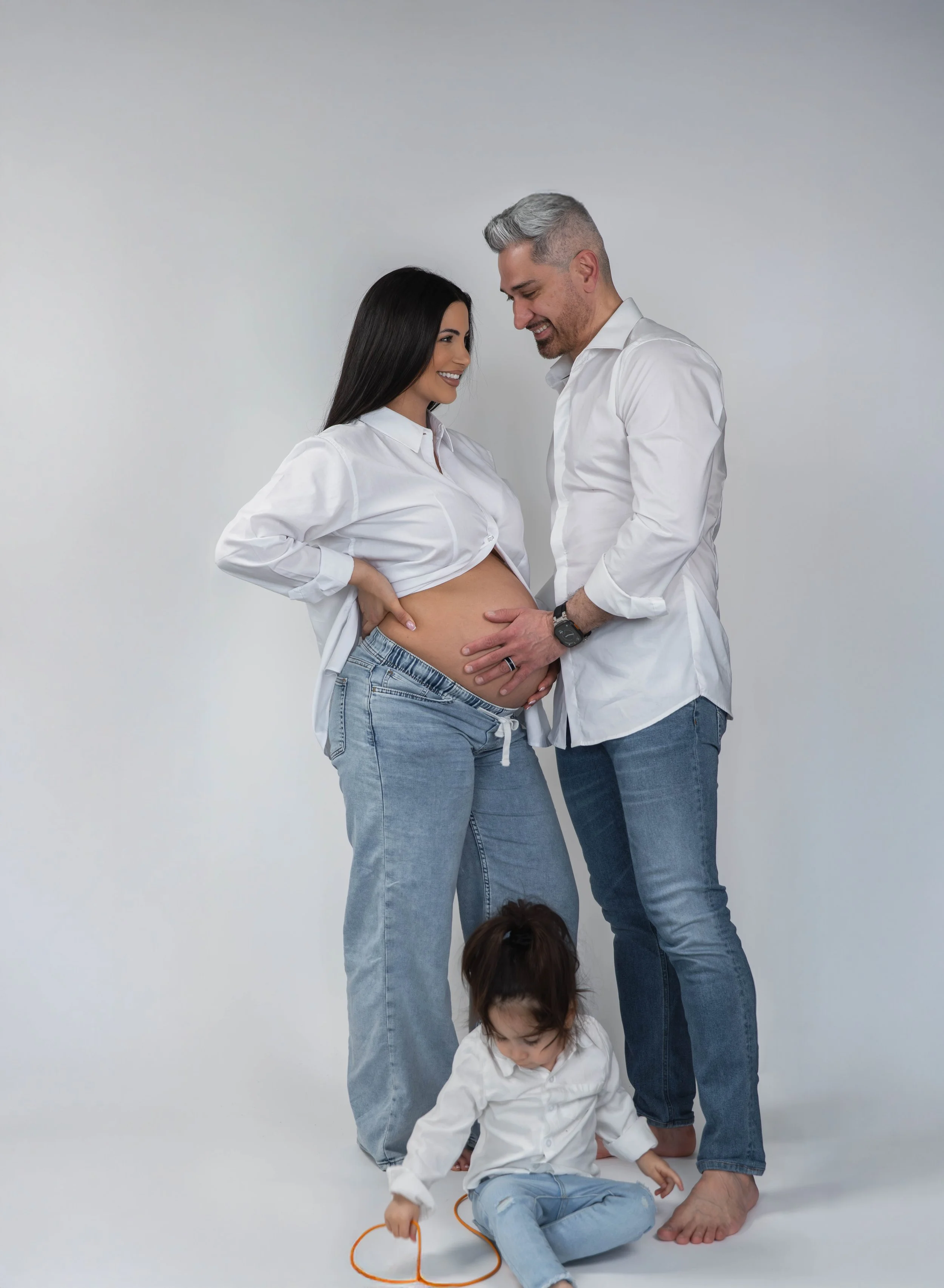 A pregnant woman with long black hair wearing a white shirt and jeans stands smiling at a man with gray hair, wearing a white shirt and jeans, who is touching her pregnant belly. Two children, a girl sitting on the floor holding an orange heart-shape