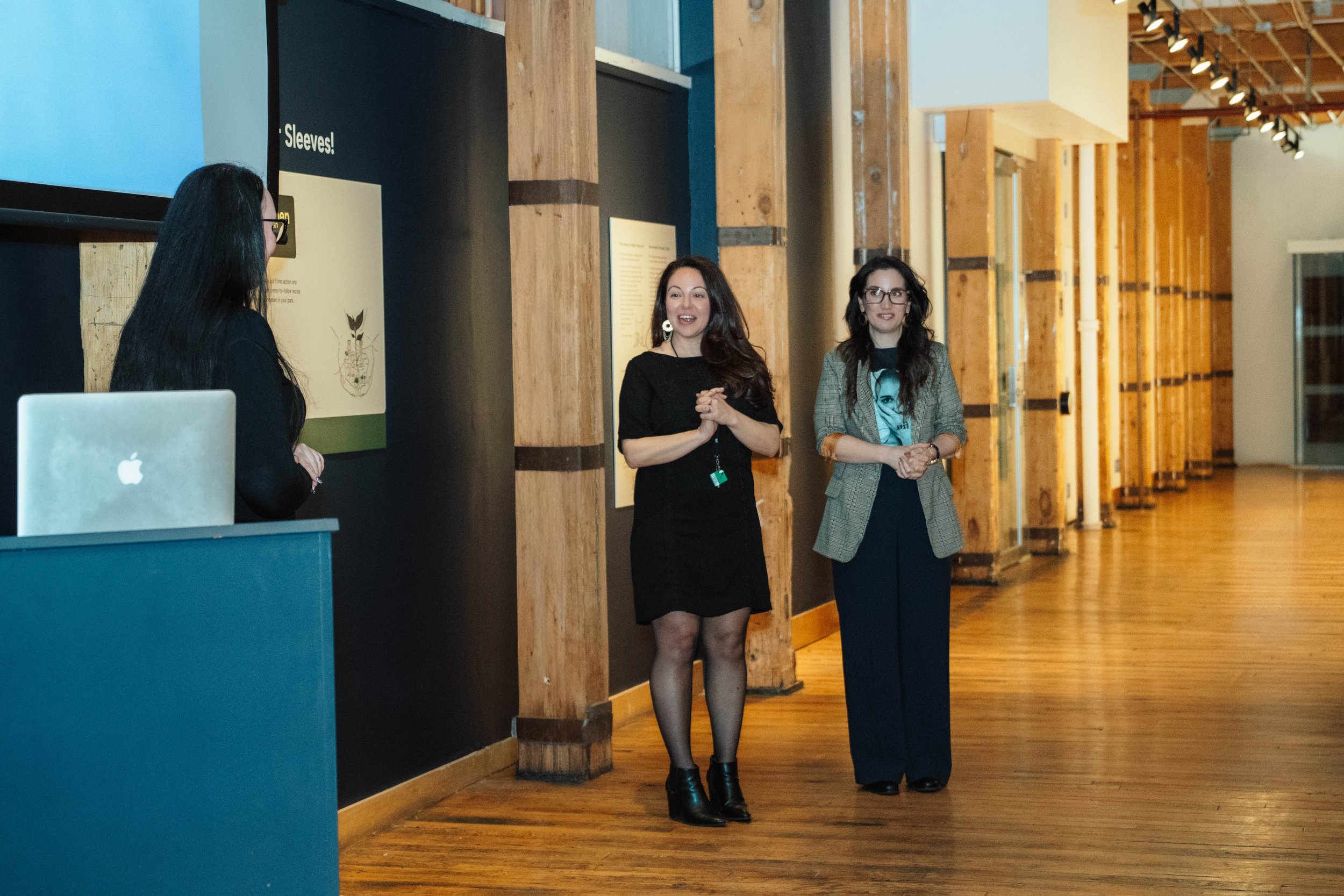 Three women are standing and talking in a long, wooden-floored room with wooden beams. One woman on the left is wearing a black dress, and the two women on the right are dressed in black and gray, with one wearing a blazer and glasses.