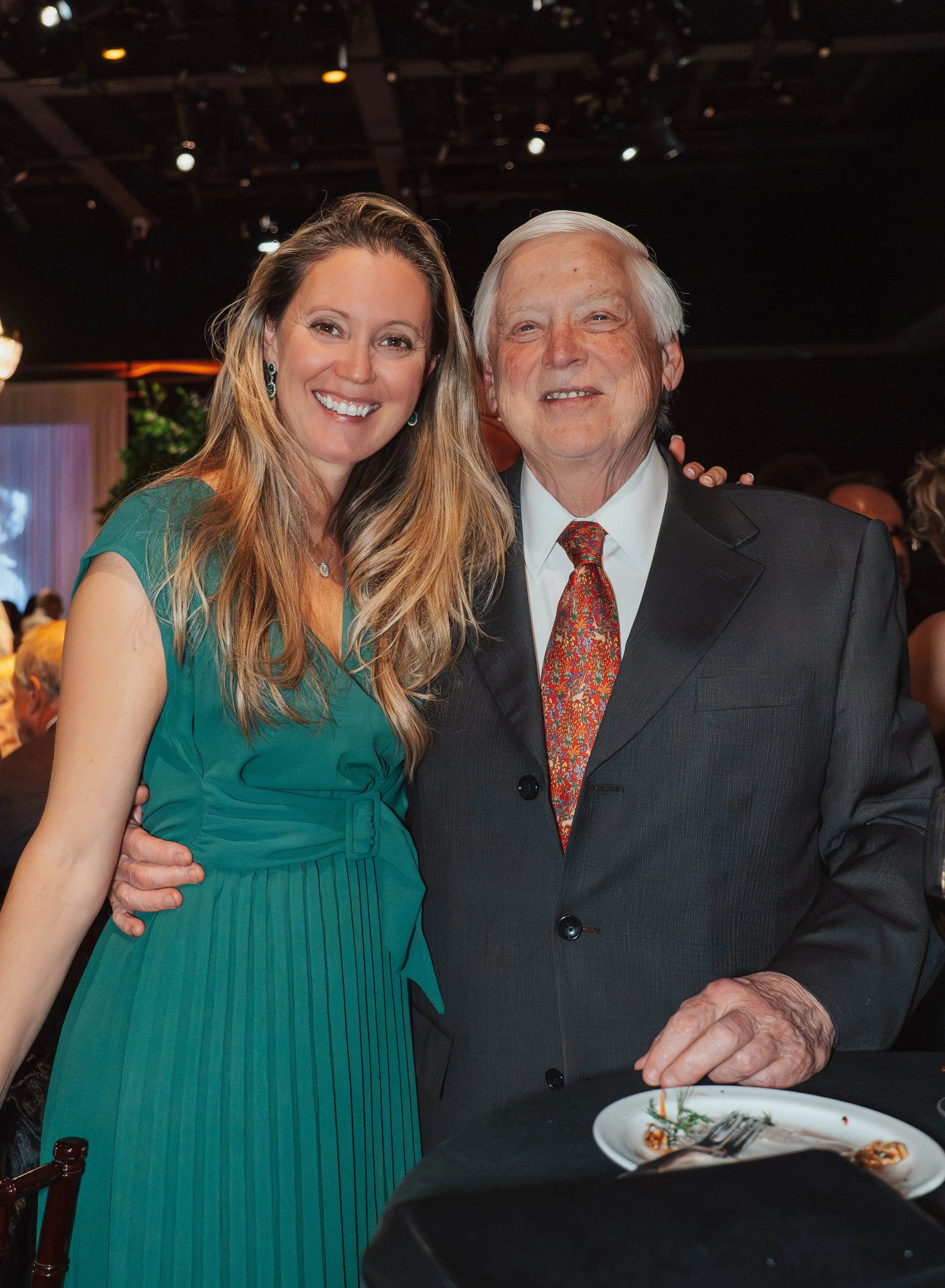 A woman and an older man in formal attire smiling together at an event, standing close with a black table with a white plate and utensils in front of them.
