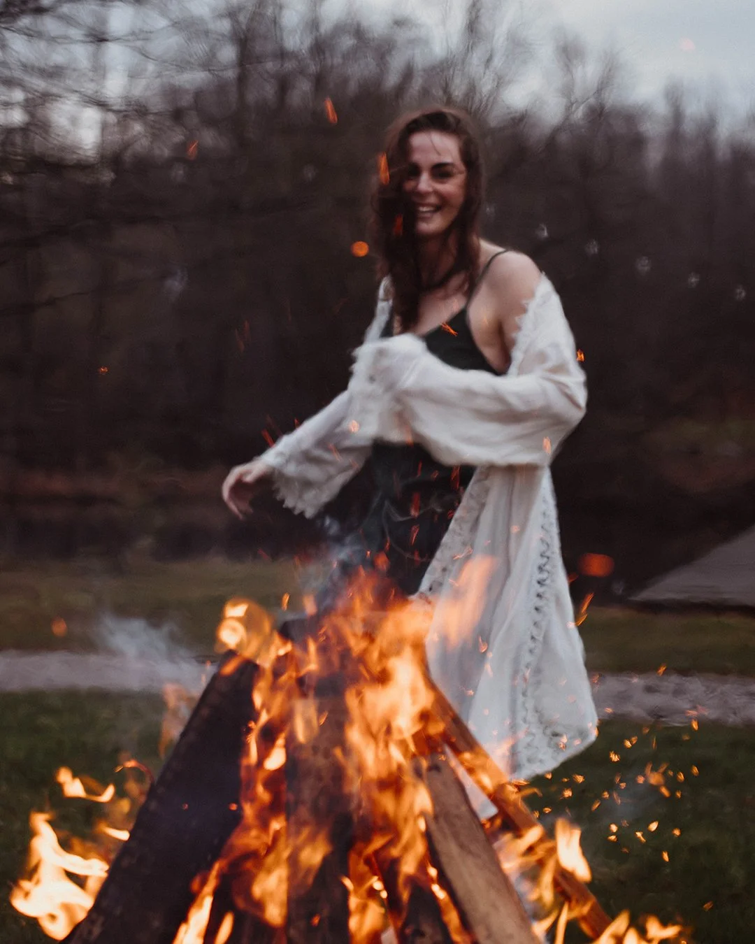 A woman with brown hair smiling while standing near a campfire outdoors during dusk.