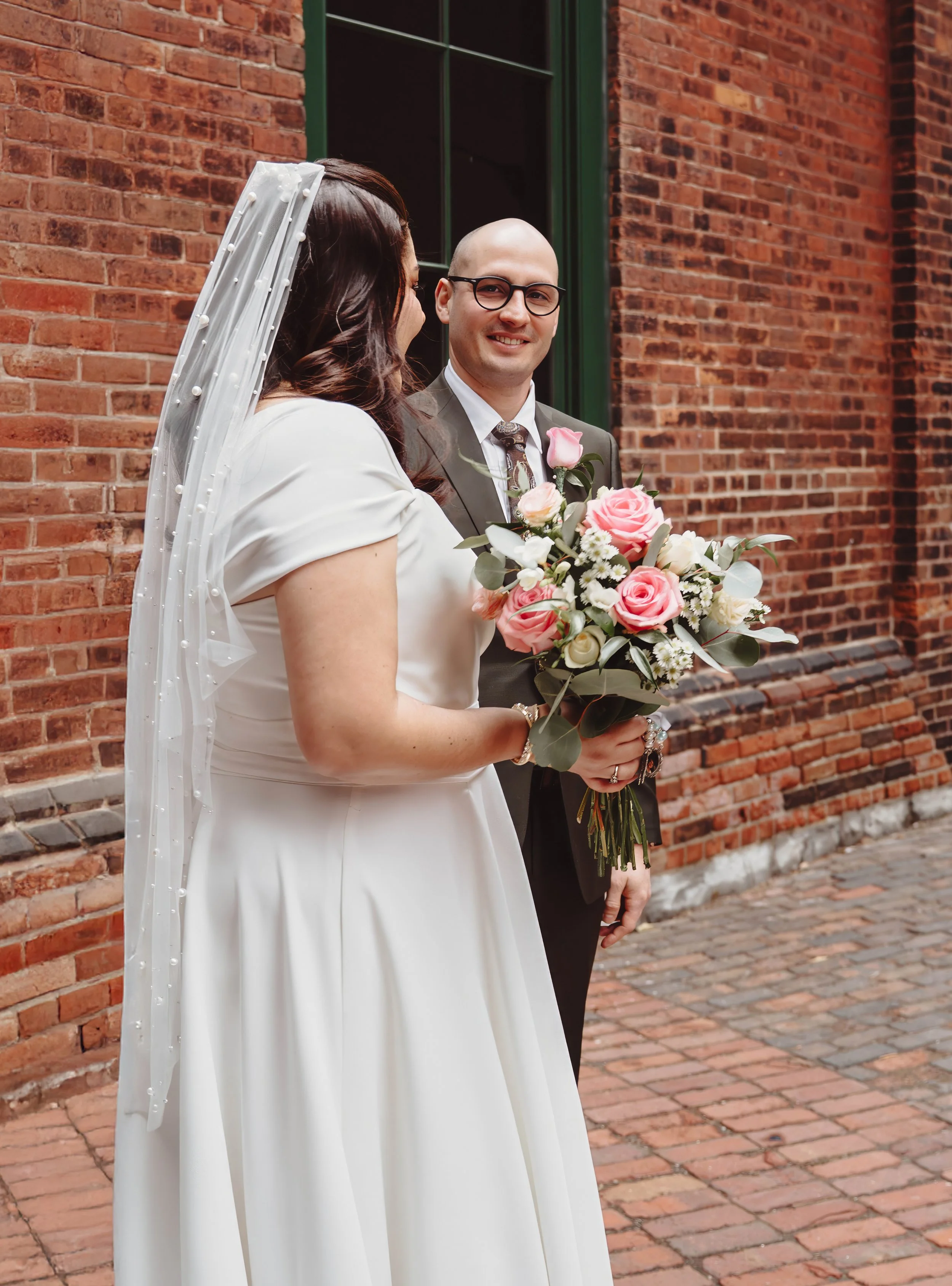 A bride and groom standing outside near a brick wall, with the bride holding a bouquet of pink and white roses.