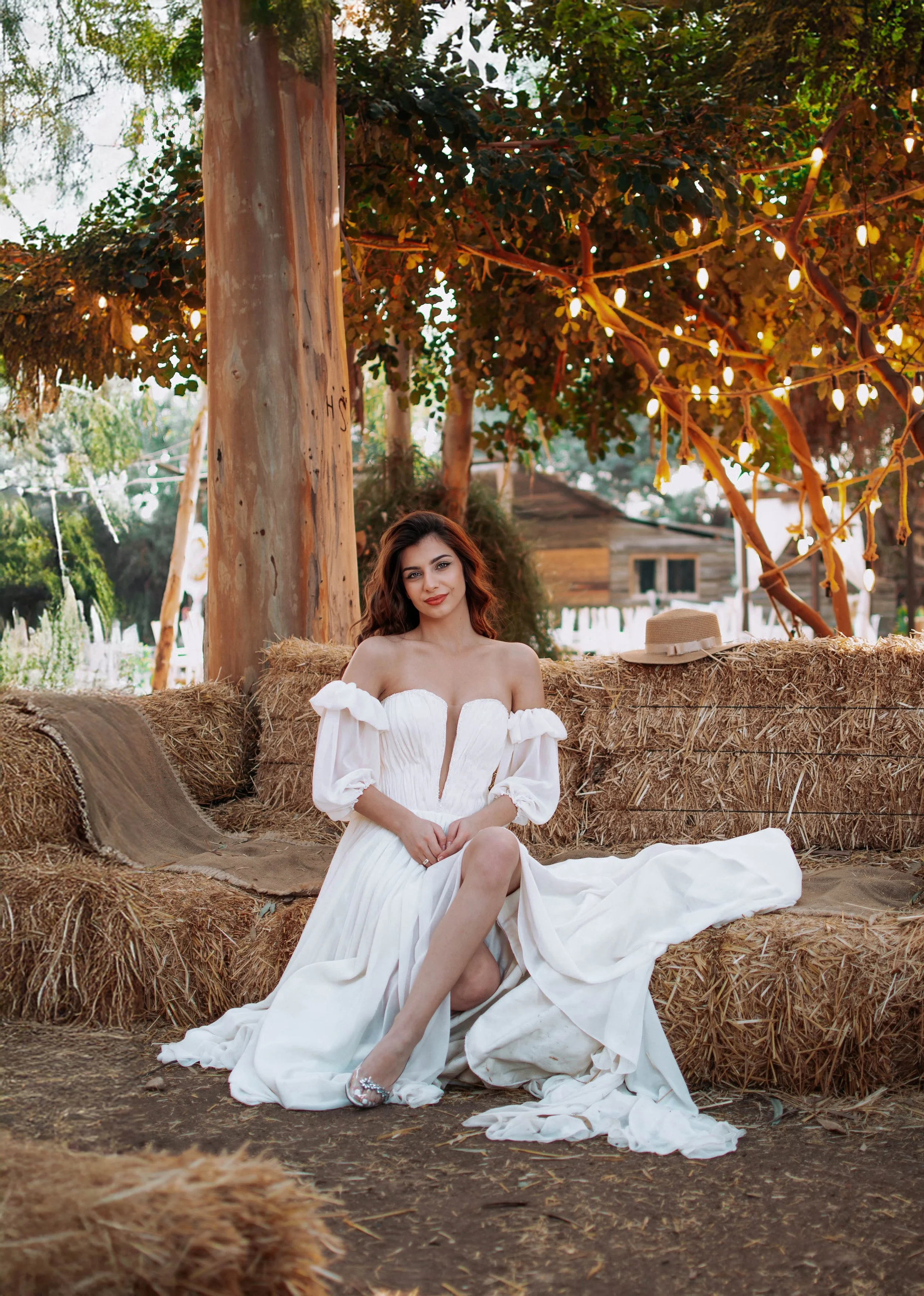 A woman with long wavy hair, wearing a white off-the-shoulder dress, is sitting on a bale of hay in an outdoor rustic setting. There are hay bales and a straw hat next to her, with trees and string lights hanging above in the background.