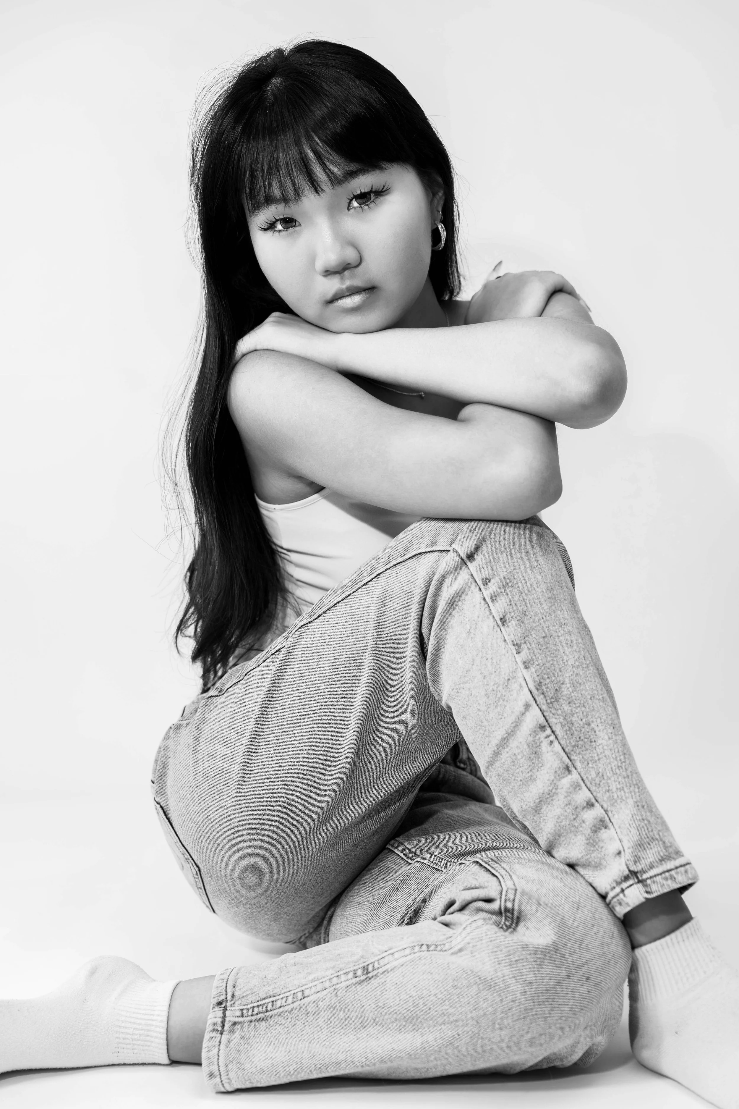 Black and white studio portrait of Dana seated against a white backdrop, wearing a white tank top and jeans with her arms resting across her knees, photographed by Lara Grace Creative Studio in Toronto.