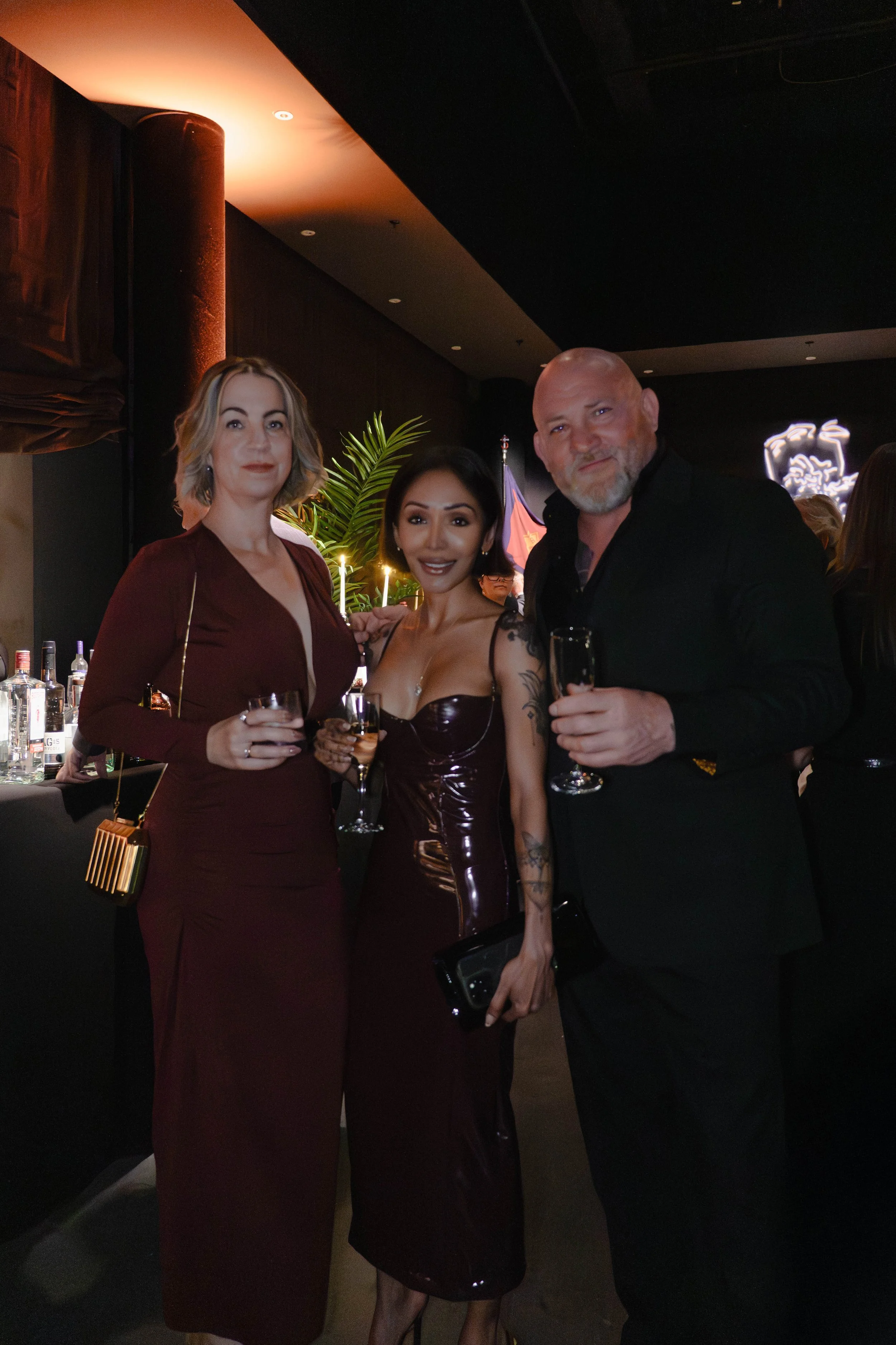 Three adults in formal attire holding glasses at a party or event, with a bar, drinks, and decorative lighting in the background.