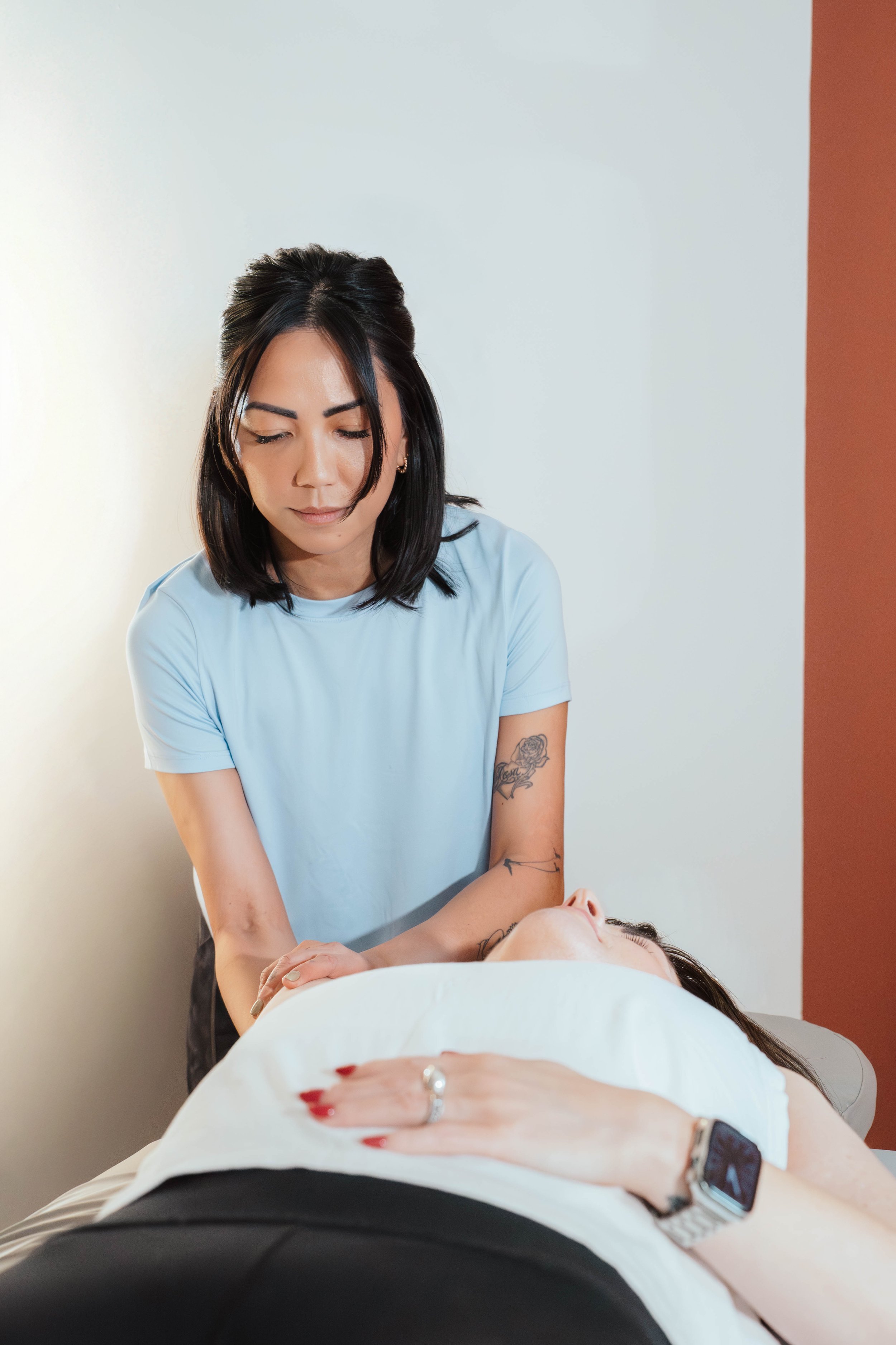 A massage therapist gives a shoulder massage to a woman lying face up on a massage table in a spa room.