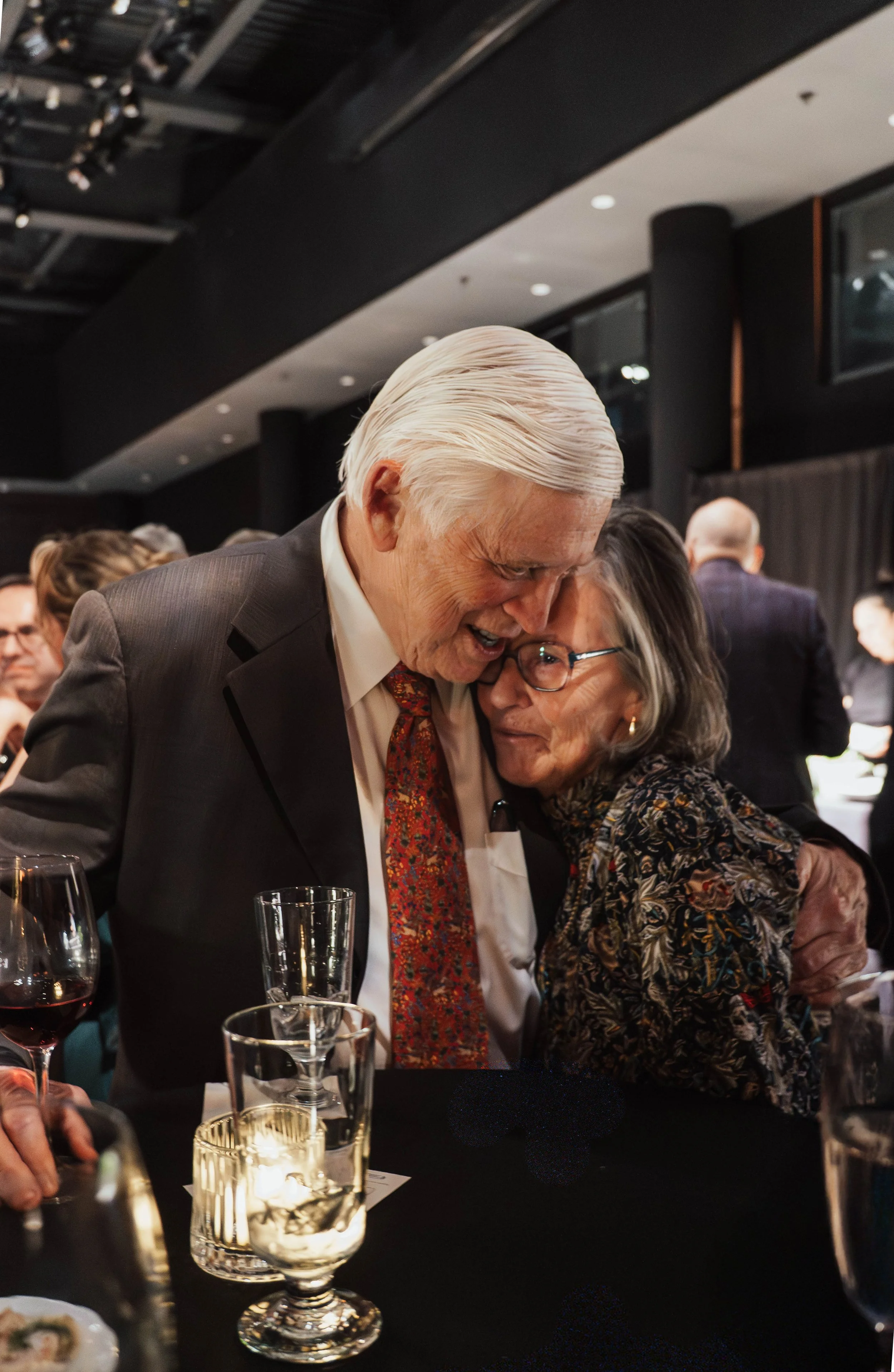 An elderly man and woman hugging at a social event, surrounded by glasses and other guests in a dimly lit room.