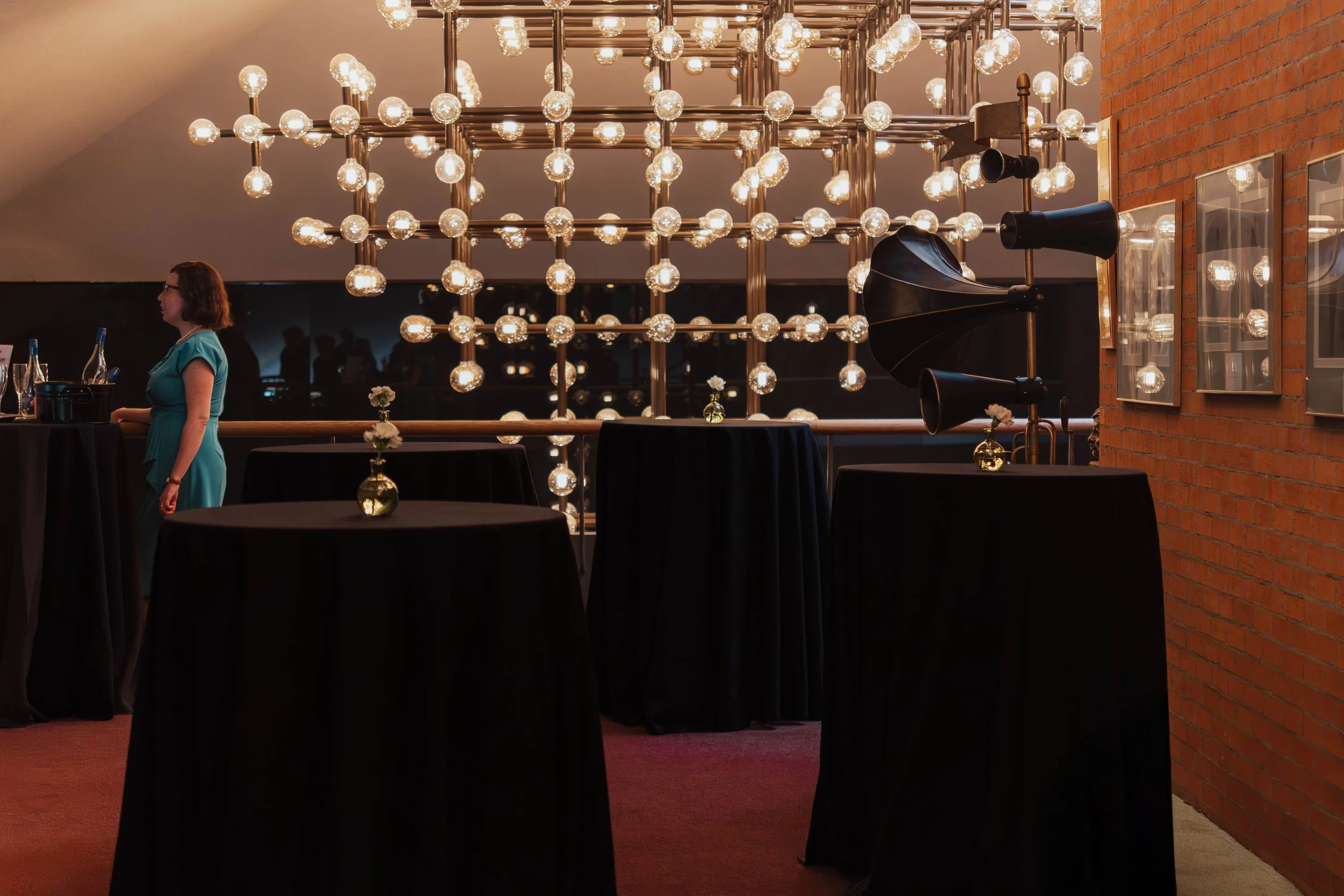 A dimly lit room with black high-top tables covered in black tablecloths, each decorated with a small glass vase holding a single white flower. A woman in a teal dress stands by a bar area on the left. A large wall of numerous exposed light bulbs cre