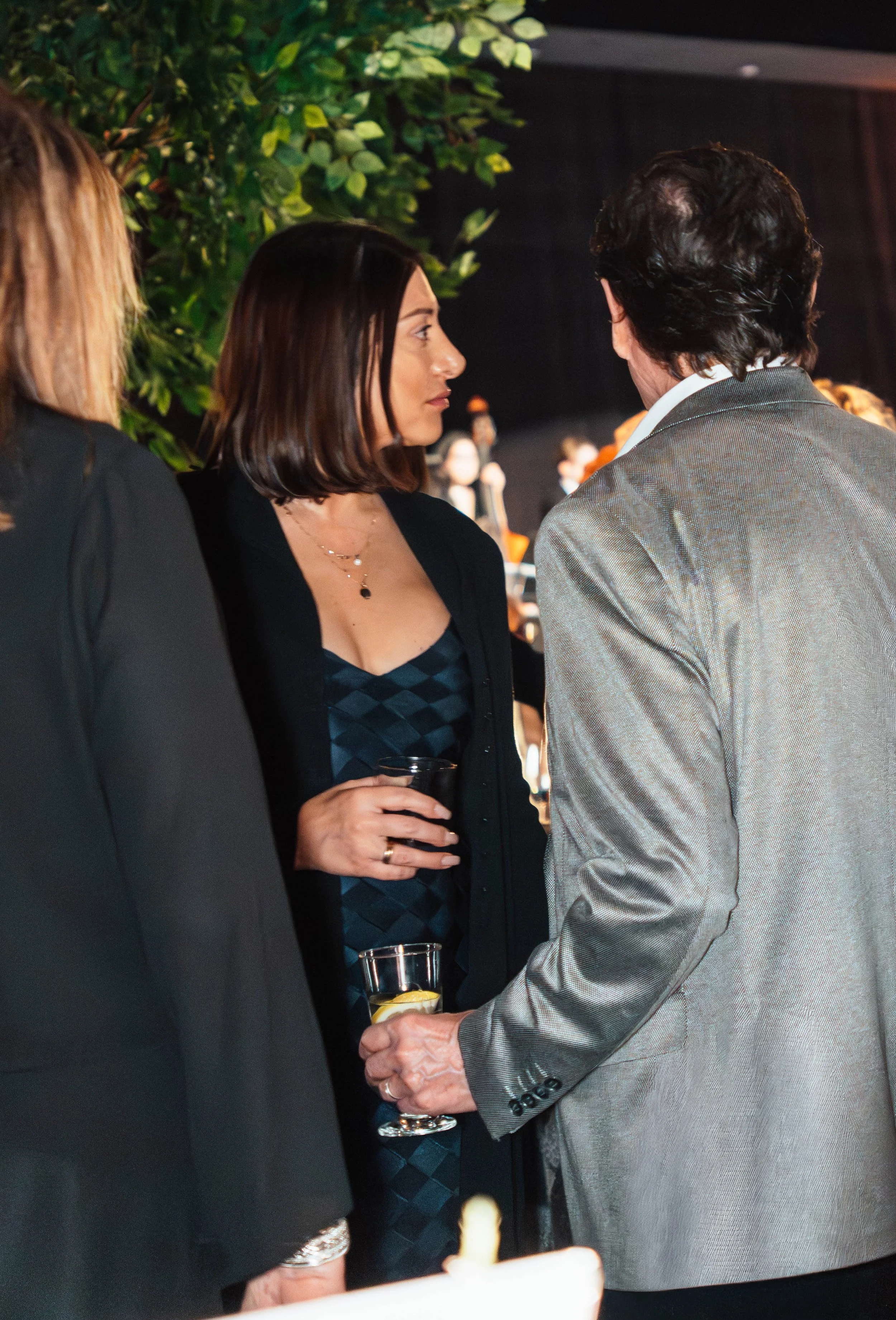 Three people engaged in conversation at an indoor event, with one woman holding a drink and wearing a black dress, a woman in black blazer, and a man in a gray suit.