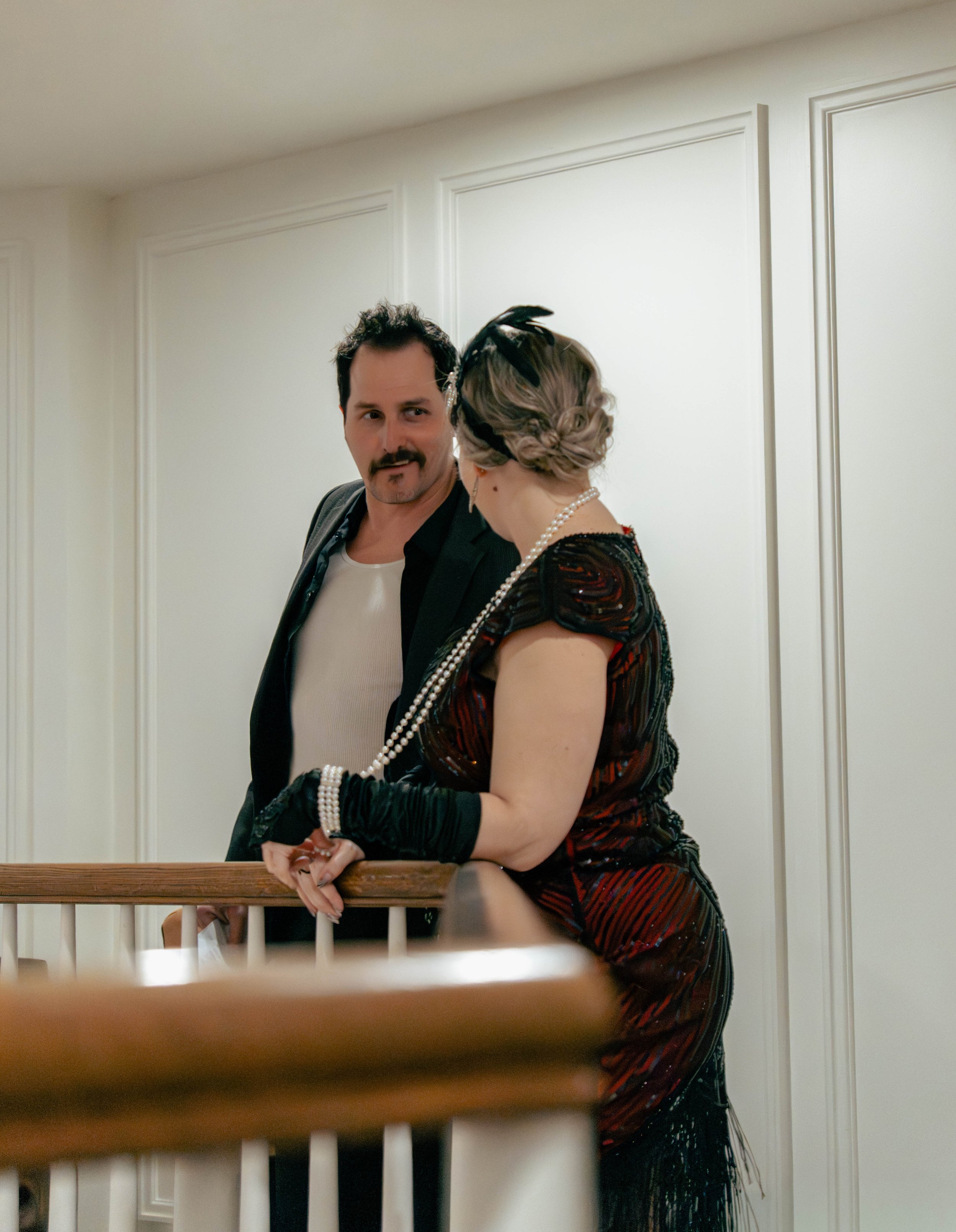A man and woman dressed in formal attire, with the woman wearing a beaded necklace and black gloves, having a conversation on a staircase in a well-lit indoor setting.