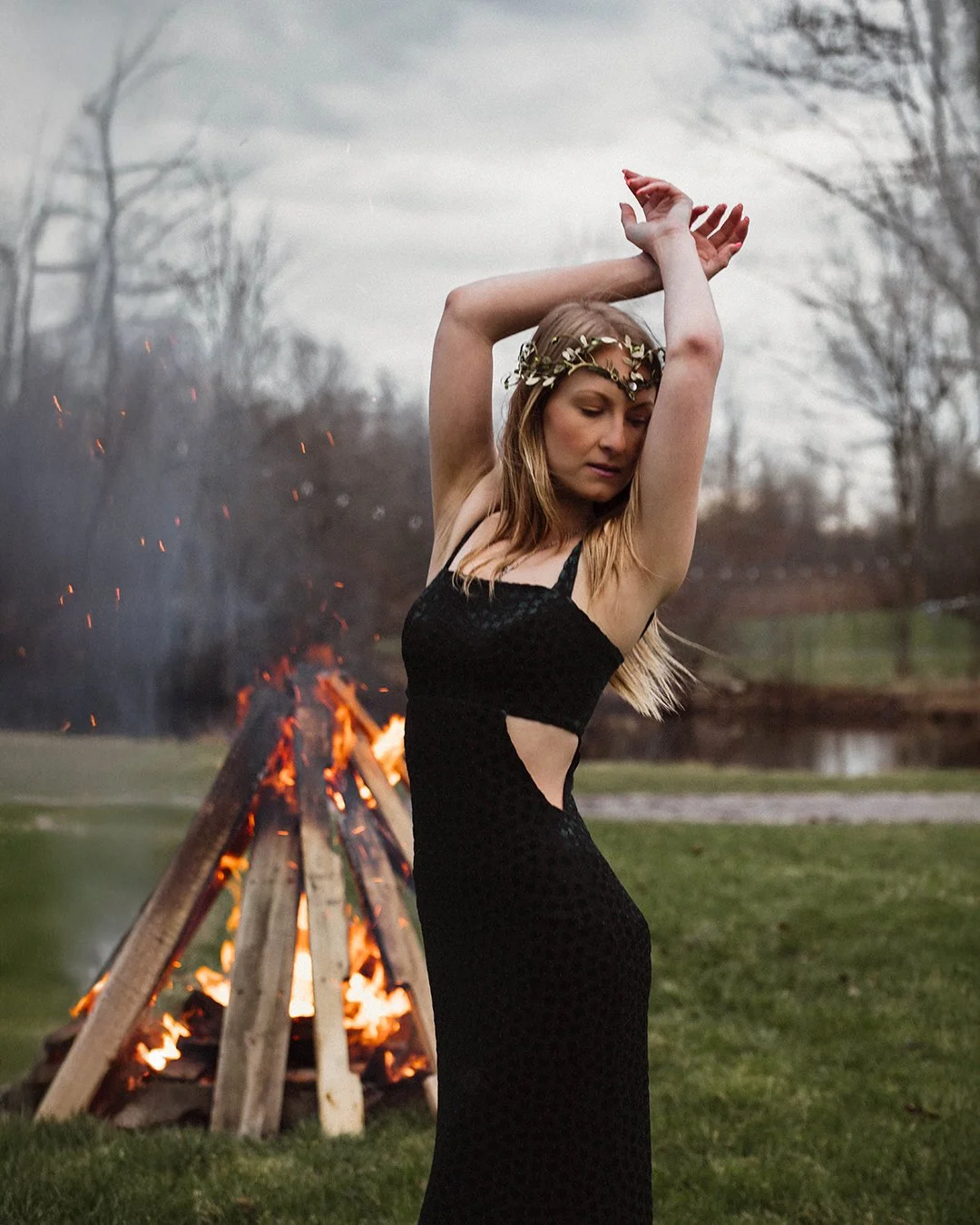 A woman in a black dress with cutouts, wearing a gold leaf headband, stands with arms raised in front of a bonfire outdoors on a cloudy day.