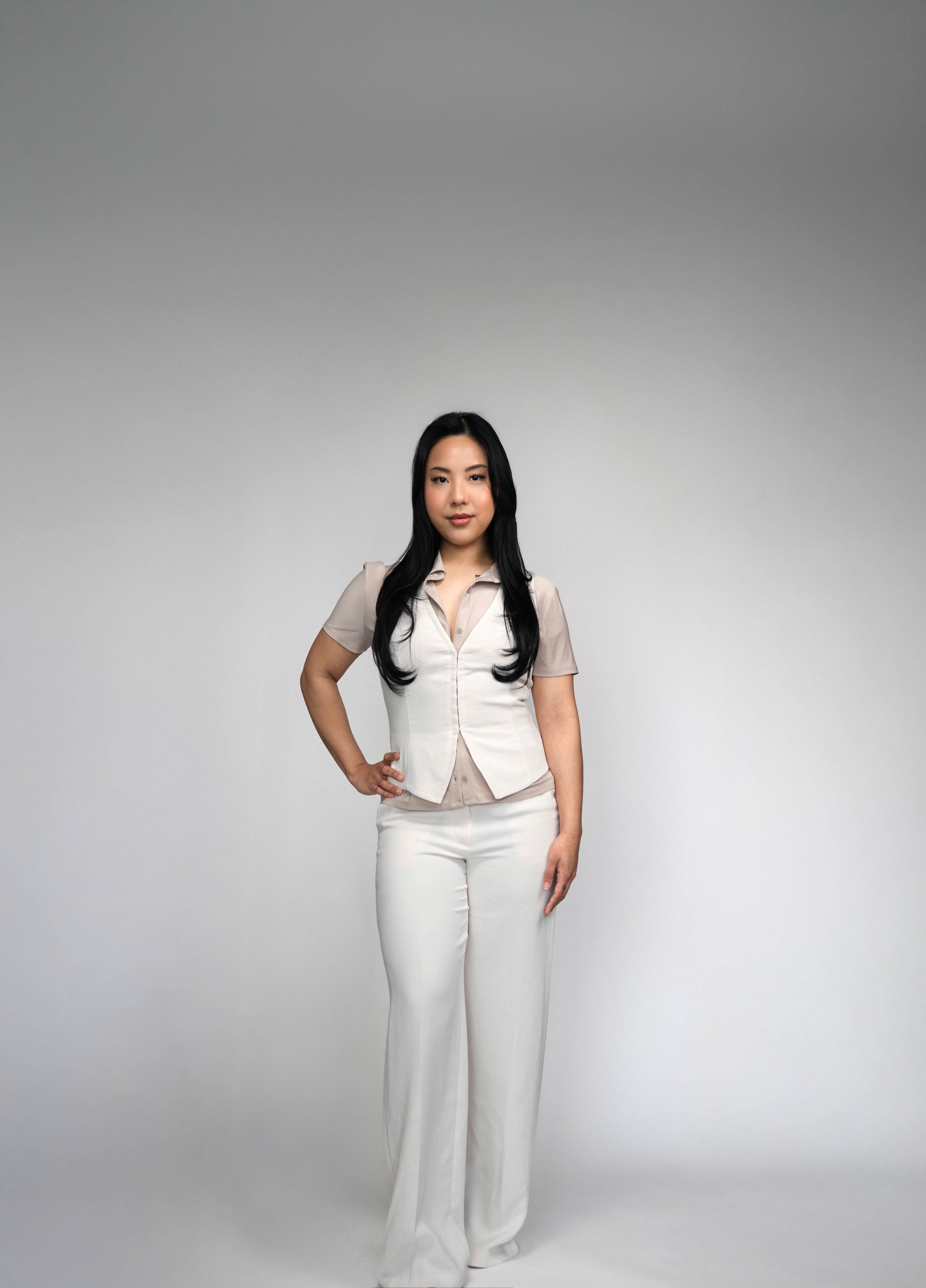 A woman with long black hair wearing a beige blouse, white vest, and white wide-leg pants standing against a plain gray background.