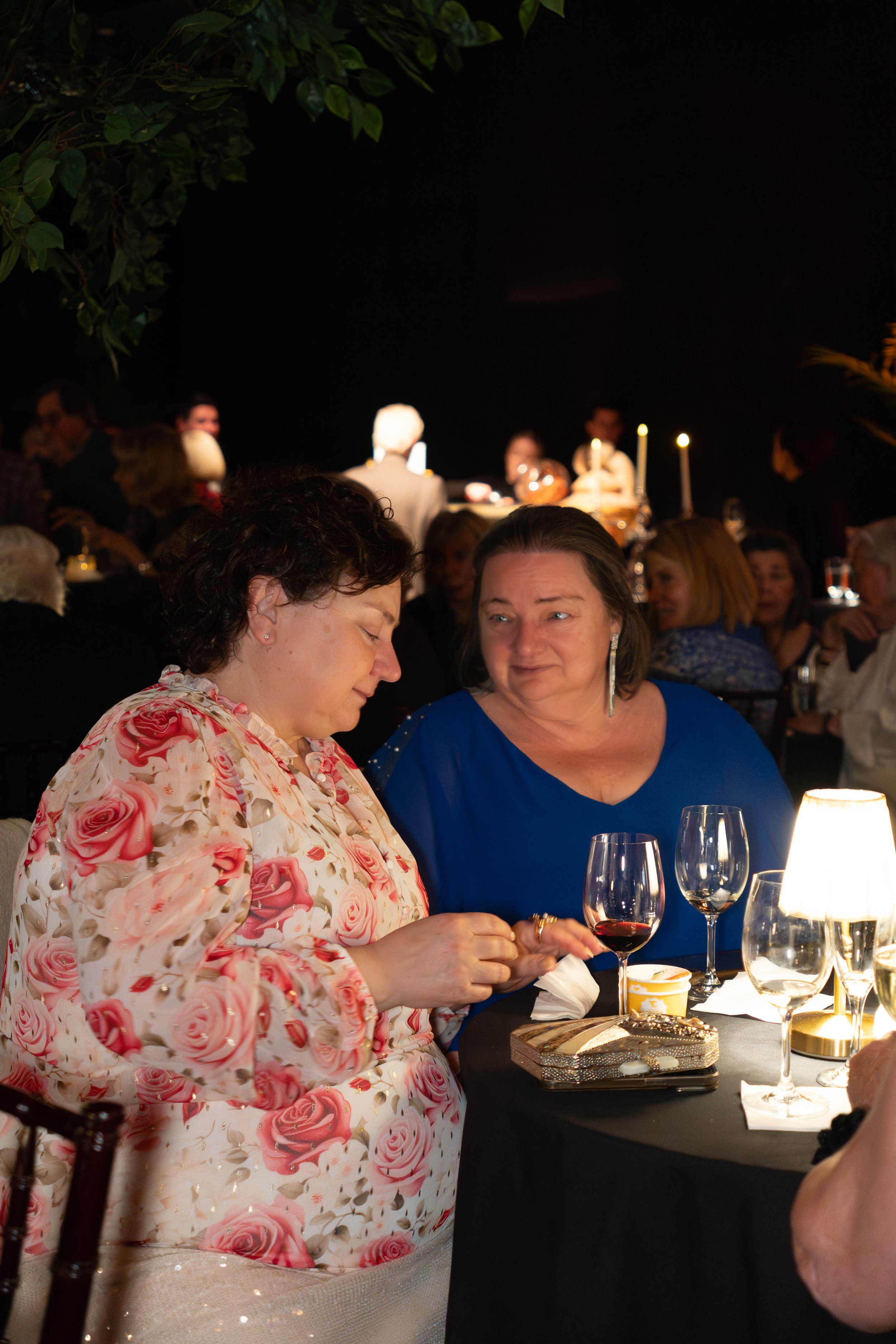 Two women sitting at a dining table, engaged in conversation, at a dimly lit social gathering or party with other people in the background.