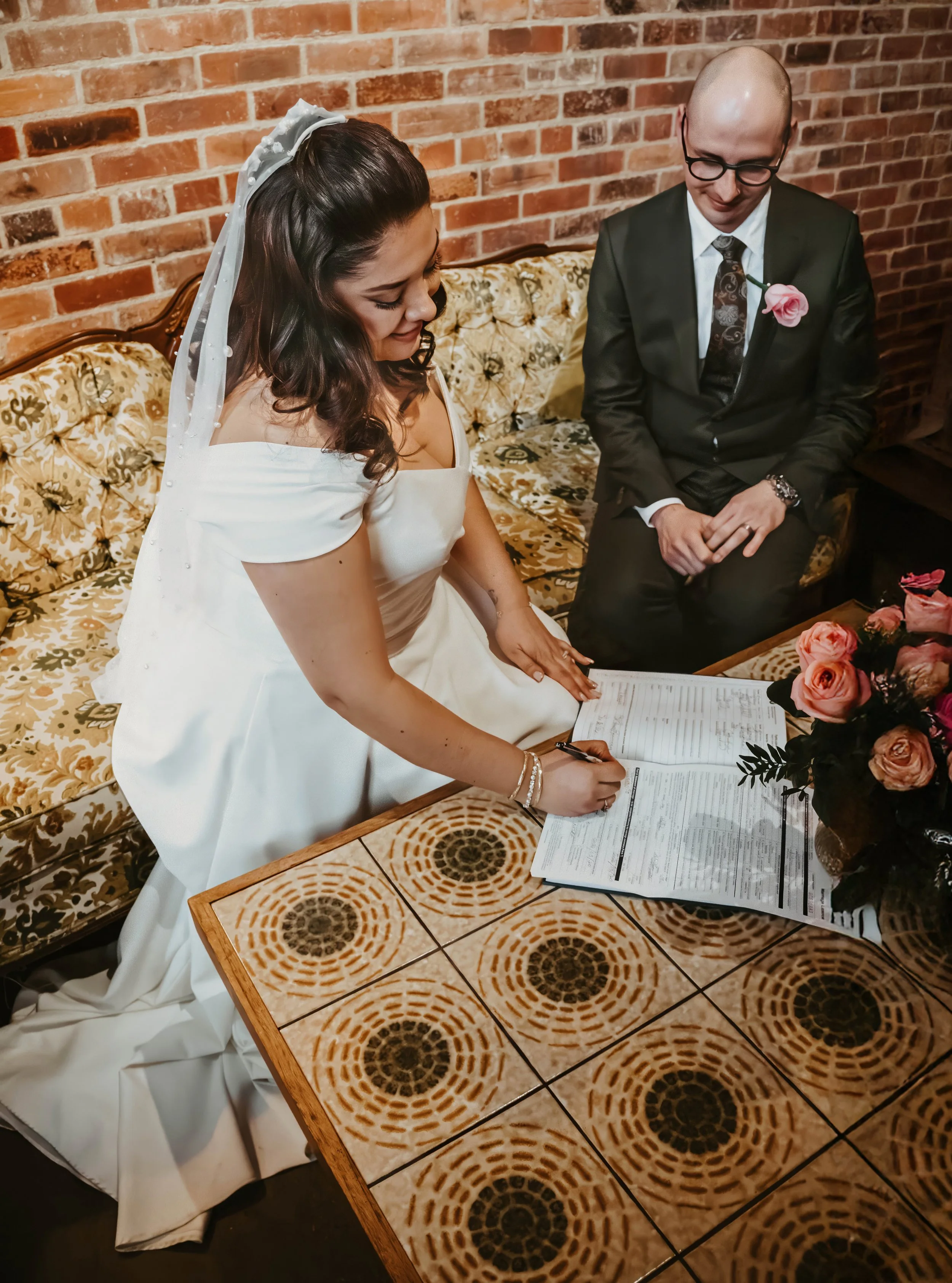 A bride signing a marriage license while sitting on a vintage floral sofa next to a groom dressed in a suit with glasses and a pink flower pinned on his lapel, in front of a brick wall and a table with a floral arrangement.