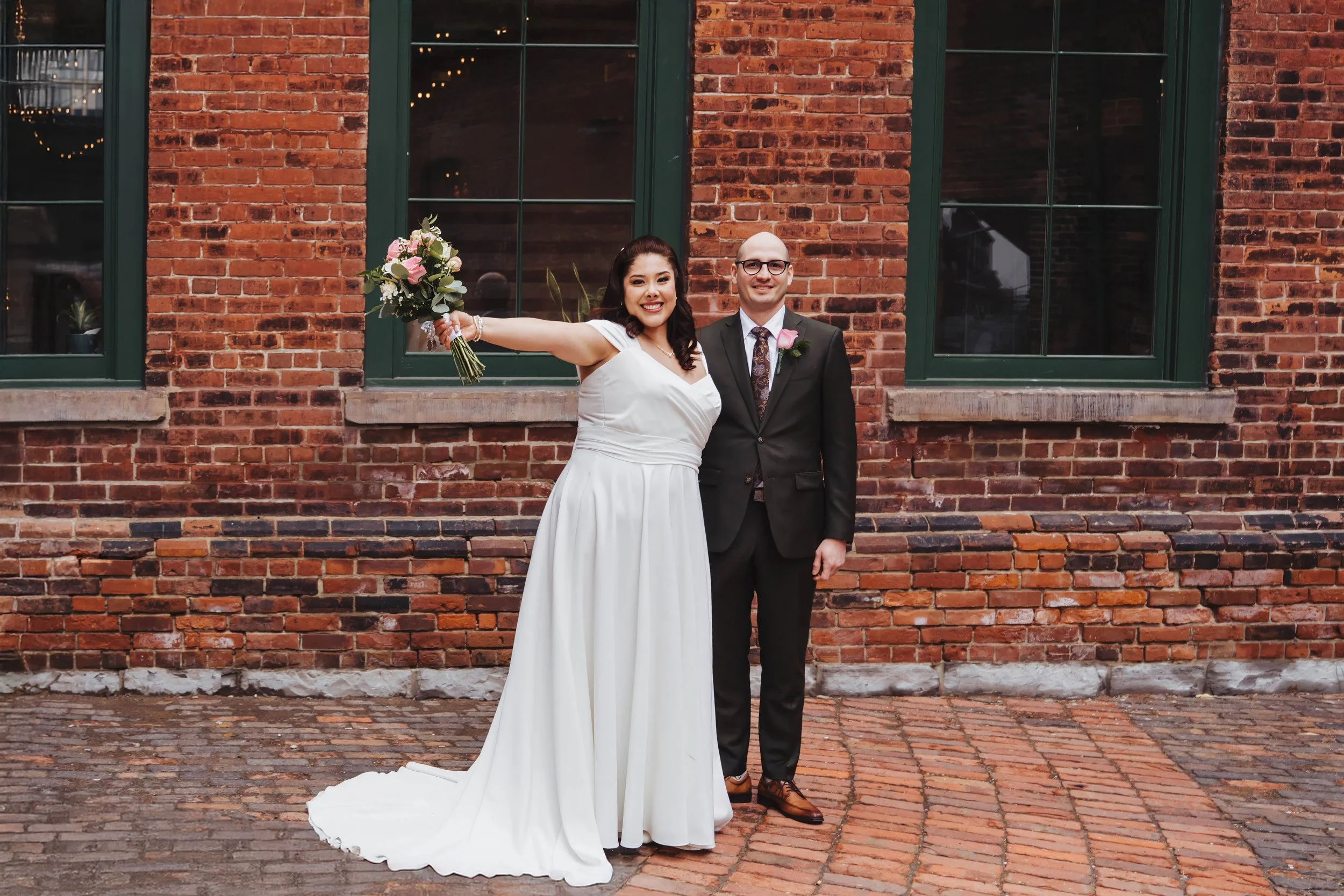 A joyful bride in a white wedding dress holding a bouquet with outstretched arm, standing beside a smiling groom in a dark suit with glasses, against a brick wall with windows.