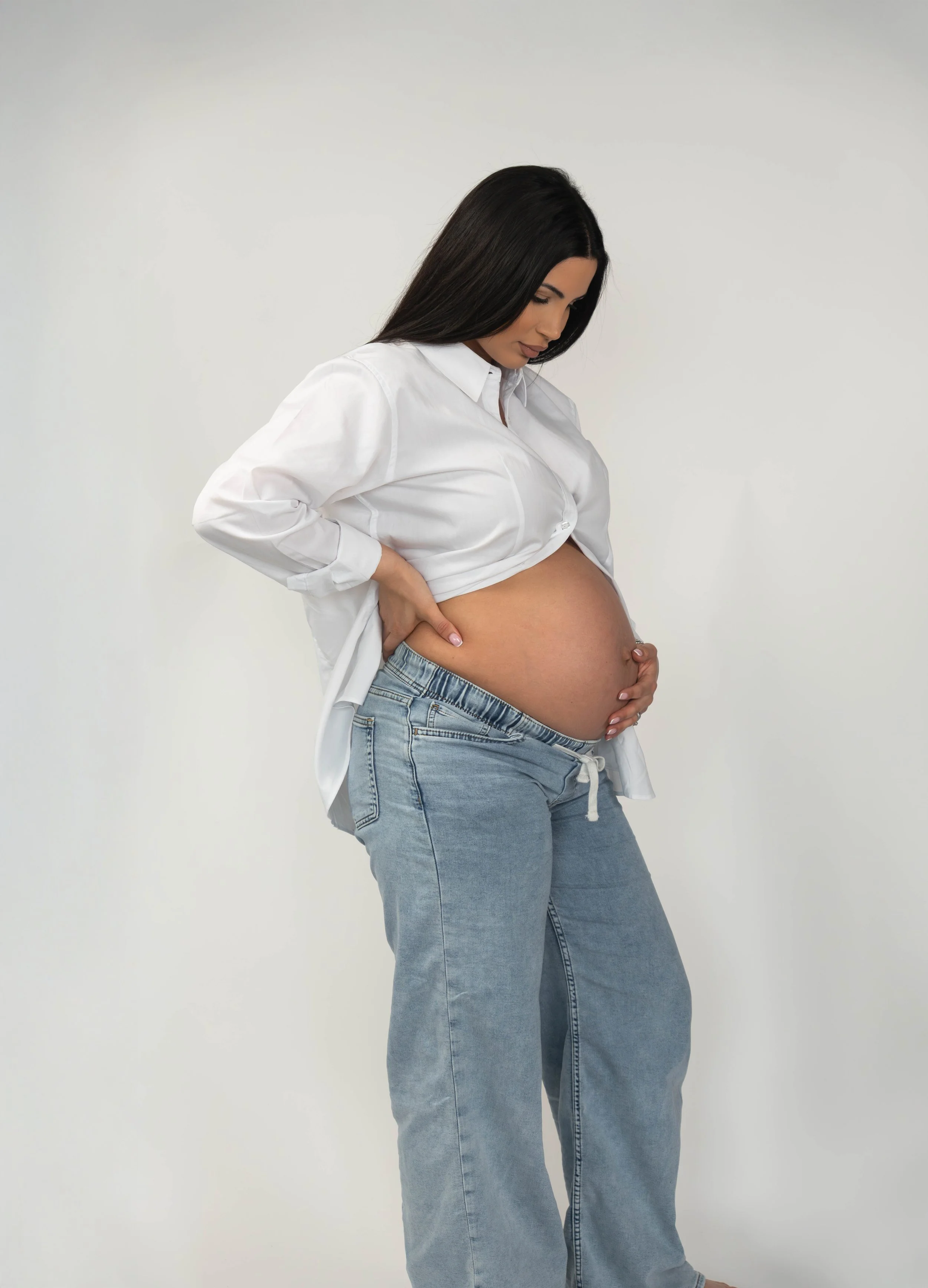 Pregnant woman with long dark hair wearing a white shirt and blue jeans, holding her belly with both hands, standing against a plain white background.