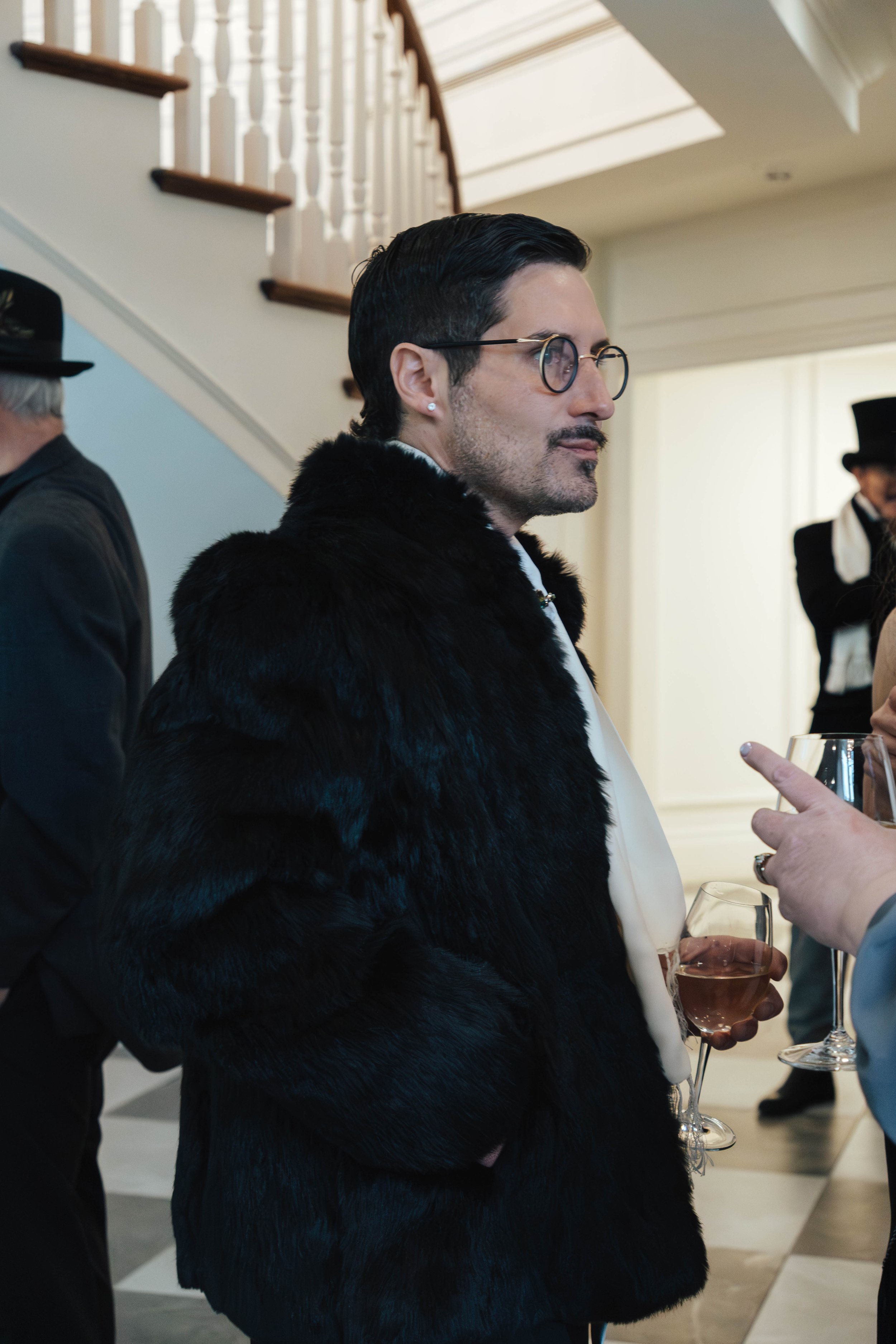 A man with dark hair, glasses, a mustache, and earrings wearing a black fur coat, holds a glass of rosé wine while socializing at an indoor event.