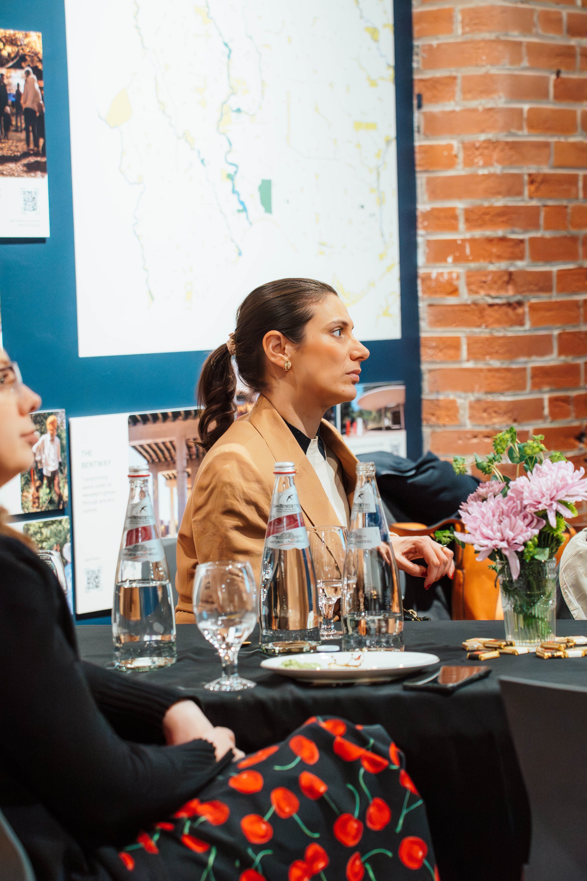 A woman with dark hair tied back, wearing a tan blazer, attending a meeting or conference, sitting at a table with water bottles, glasses, and a vase of pink flowers, with a large map on the wall behind her.