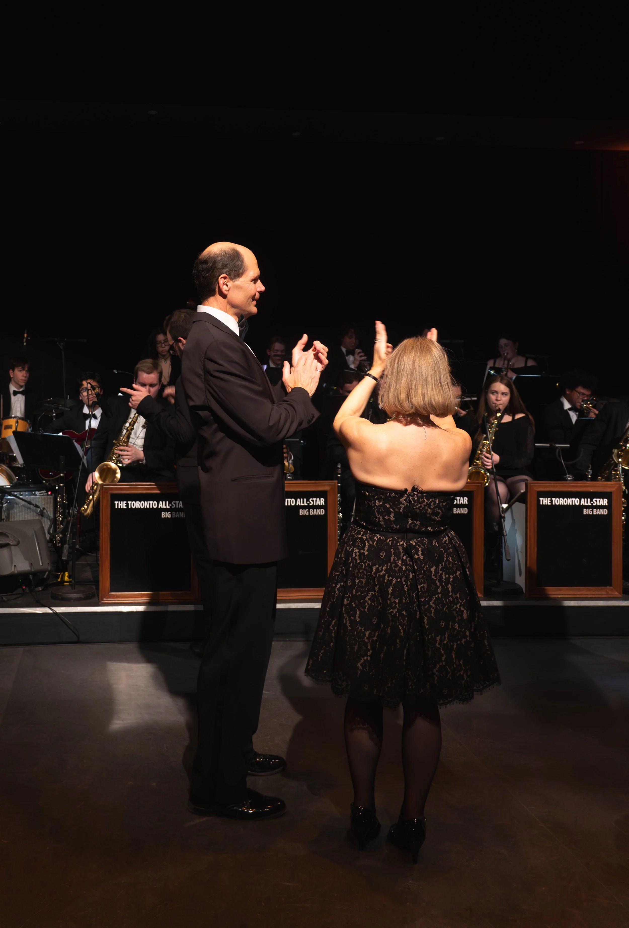 A man in a tuxedo and a woman in a black lace dress with a strapless top dance together in front of a jazz band on stage. The band members are wearing black and white formal attire, and their instruments are visible, including saxophones and drums. T
